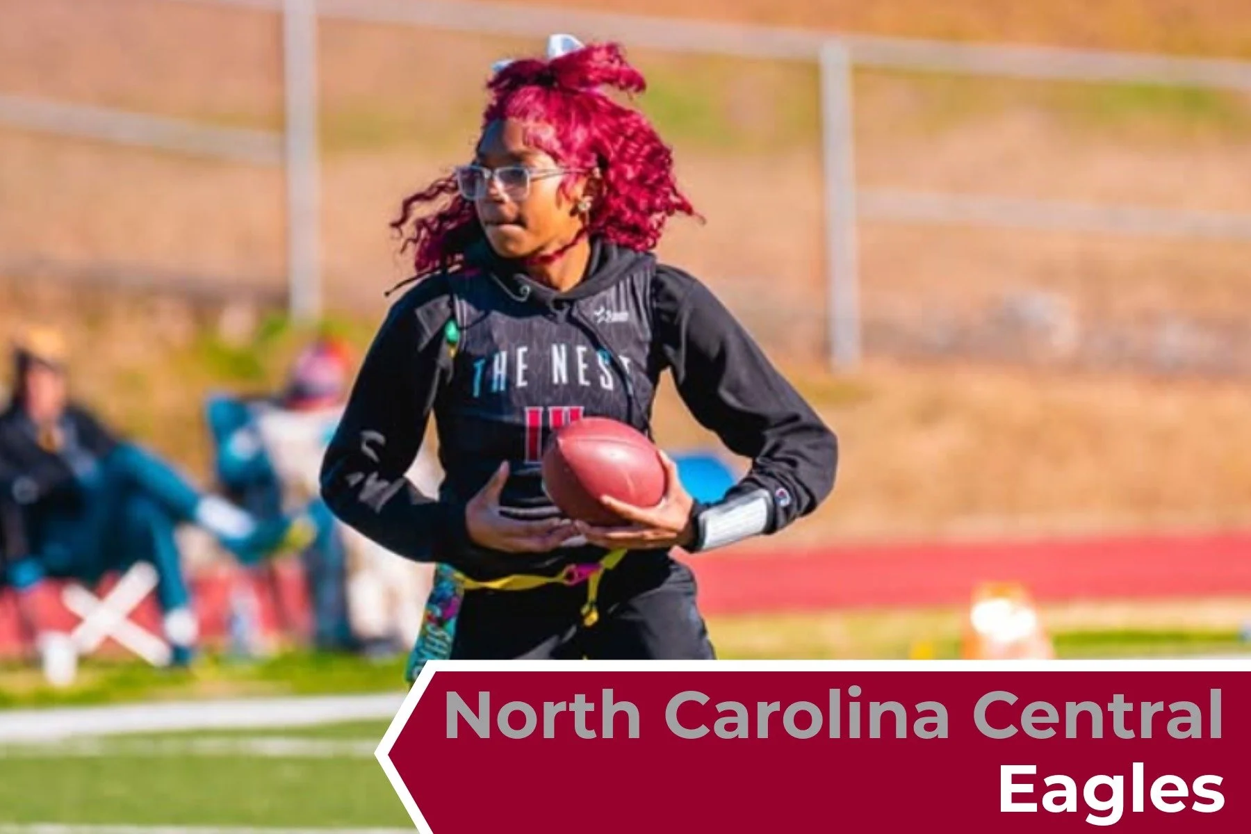 A young woman with bright pink curly hair, wearing glasses and a black sports jacket, is running on a field while holding a football. There are blurred people sitting on the grass in the background, and a sign in the bottom right corner reads "North 