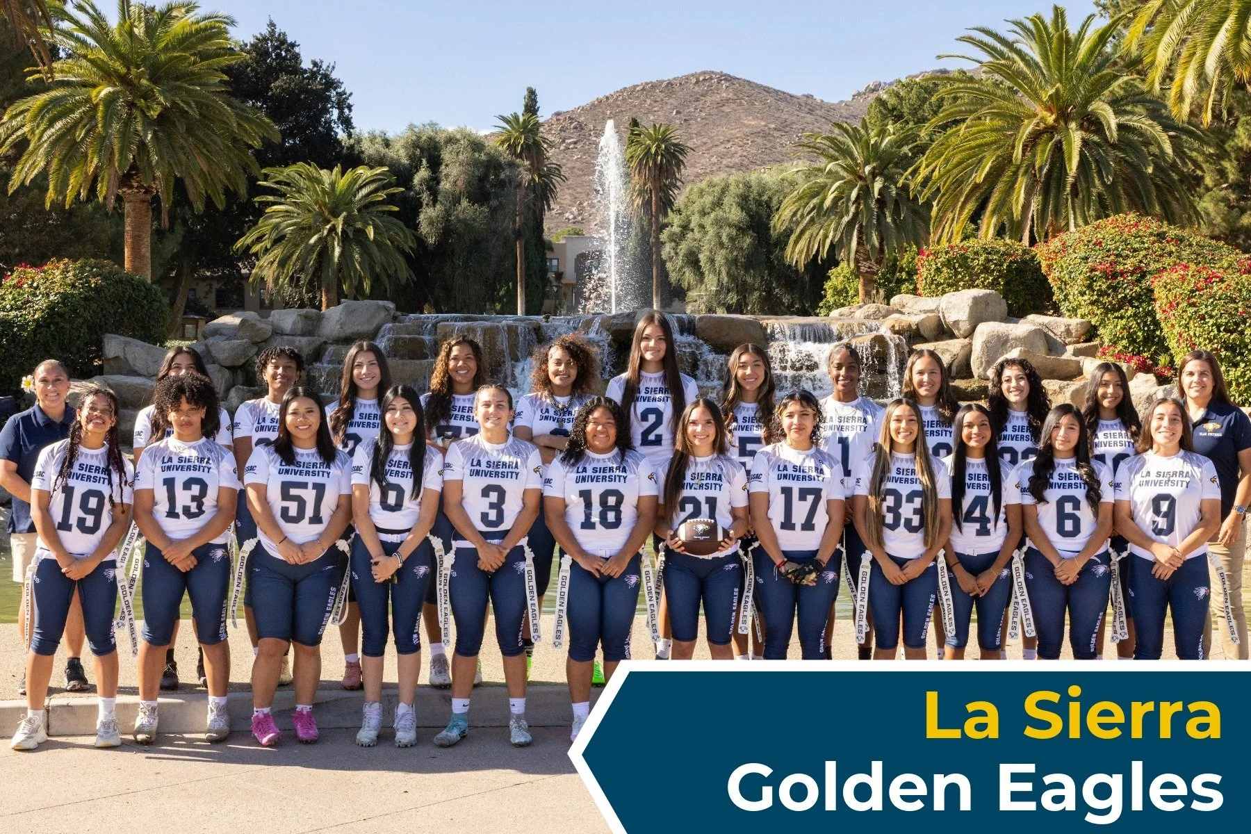 A group of female football players from La Sierra University Golden Eagles team posing outdoors in front of a fountain and palm trees, with mountains in the background, during daytime.