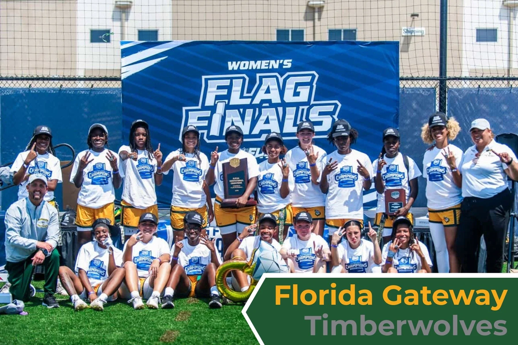 A group of young girls in sports uniforms and coaches celebrating with trophies after winning a women's flag football championship. They are posing on a sports field with a blue banner in the background that reads 'Women's Flag Finals' and a sign ind