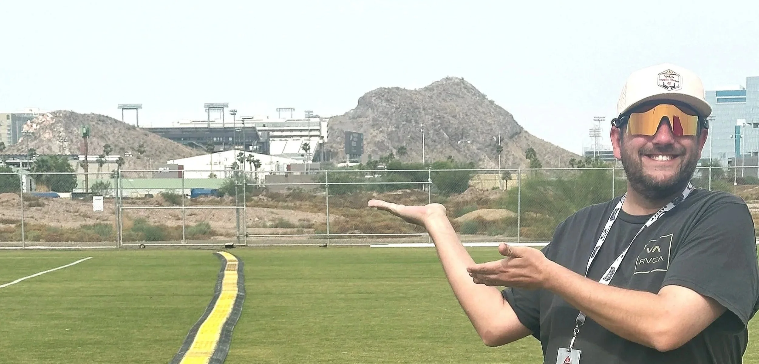 A man wearing sunglasses and a baseball cap smiling and gesturing towards a mountain landscape in the background.