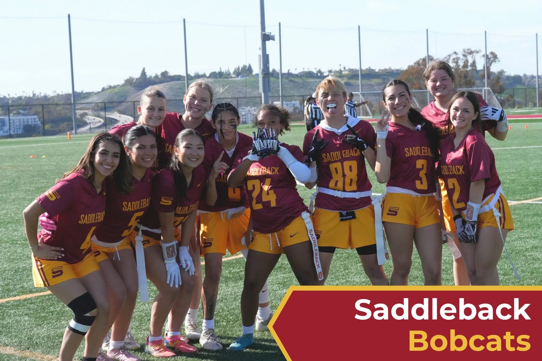 A group of girls in maroon and yellow American football uniforms posing on a football field, with a red banner at the bottom that says 'Saddleback Bobcats'.