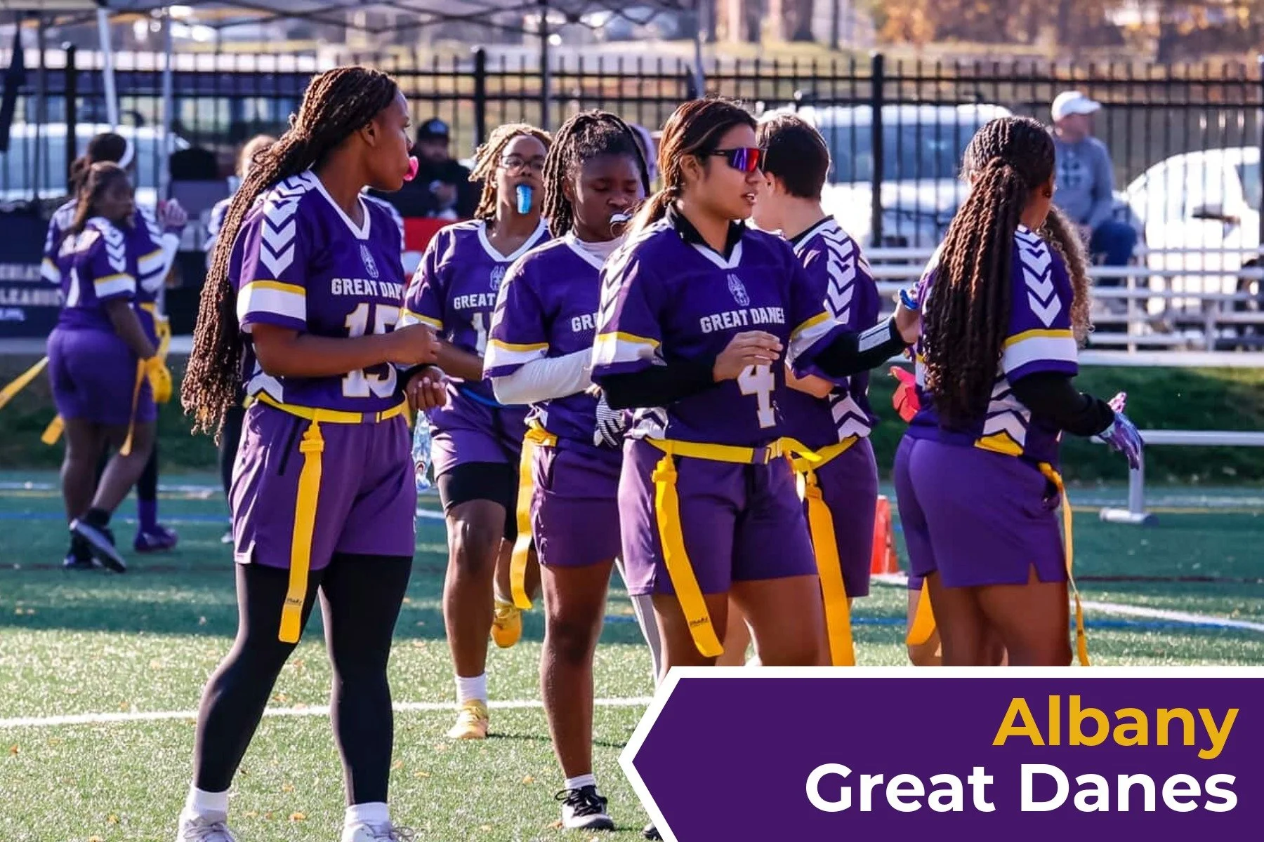A group of female athletes wearing purple and yellow uniforms with 'Great Danes' written on them, standing on a sports field during daytime, with some wearing sunglasses and carrying water bottles.