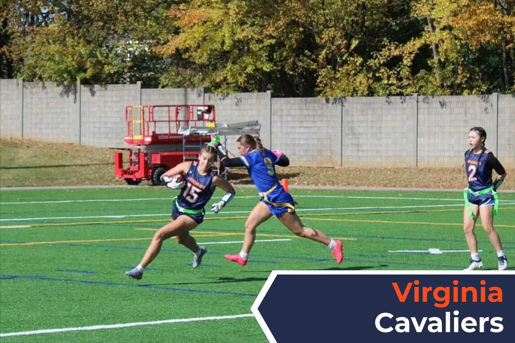 Three young female athletes play flag football on a grassy field, with trees in the background and a red fire truck parked behind a brick wall. One girl, wearing jersey number 15, is running with the football while another girl, in jersey number 5, i