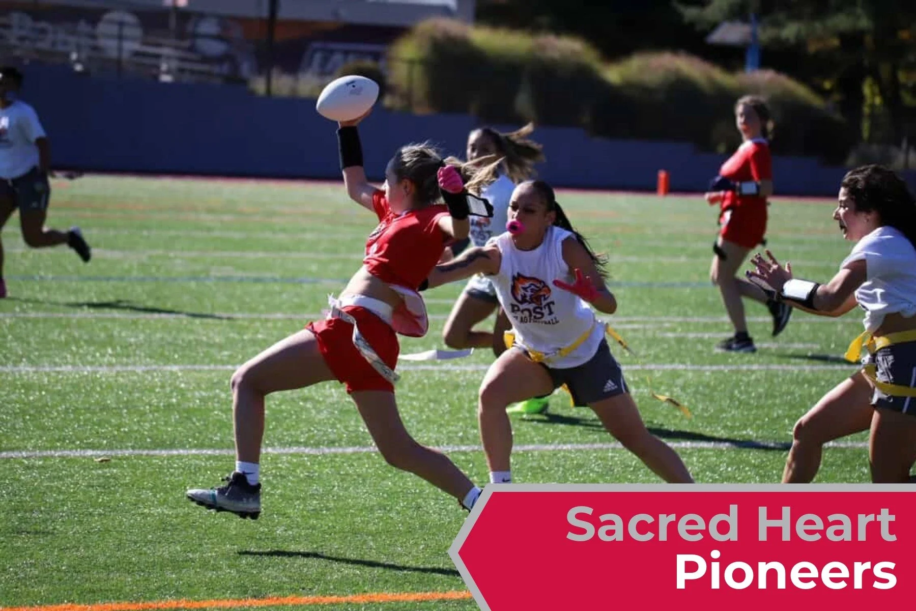 Sacred Heart University playing flag football on a grassy field, some wearing team uniforms and flags around their waists, with one girl in a red jersey jumping to catch a football, and others actively participating in the game.