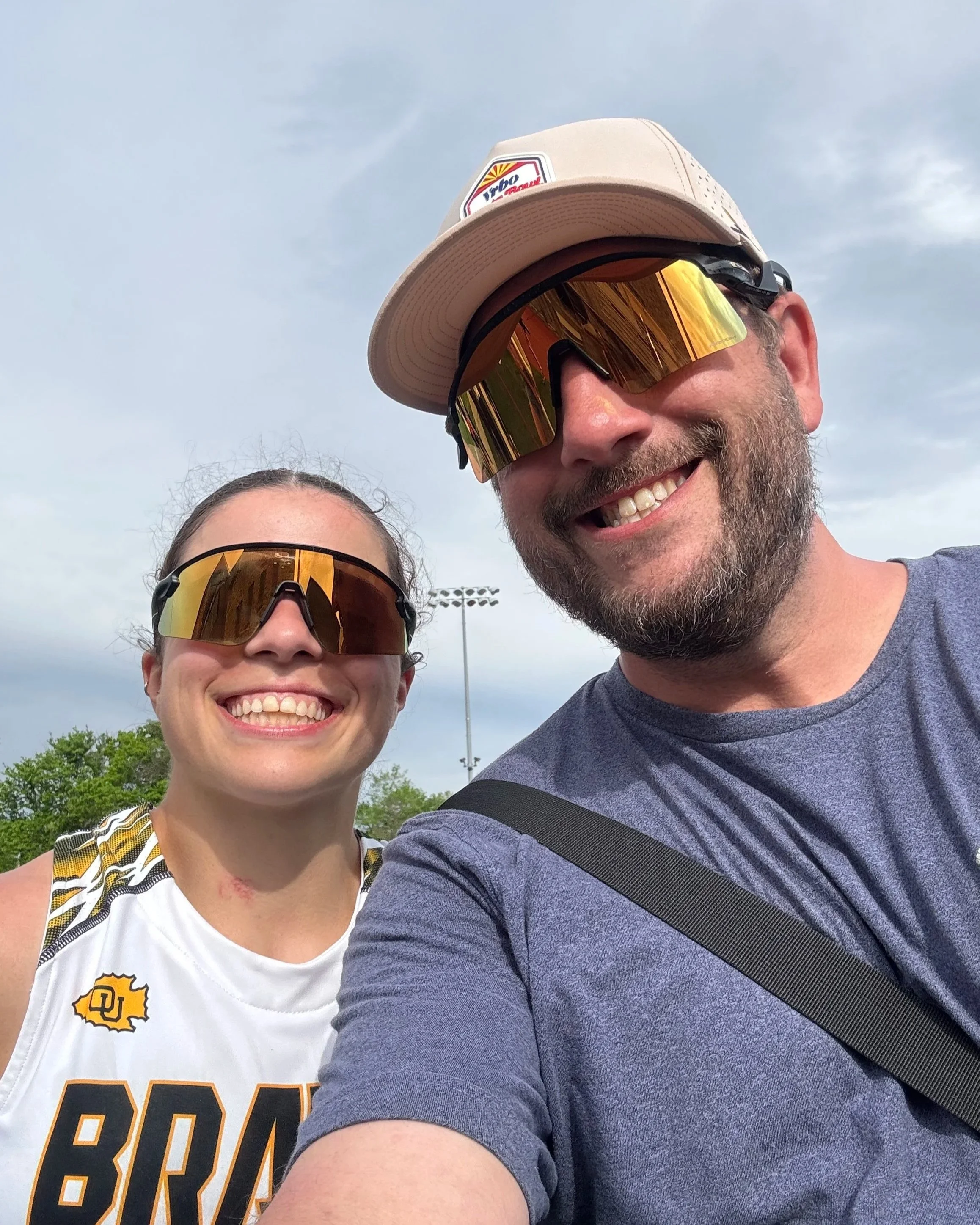 A smiling man and woman taking a selfie outdoors under a cloudy sky, both wearing sunglasses, with trees and a stadium's light poles in the background.