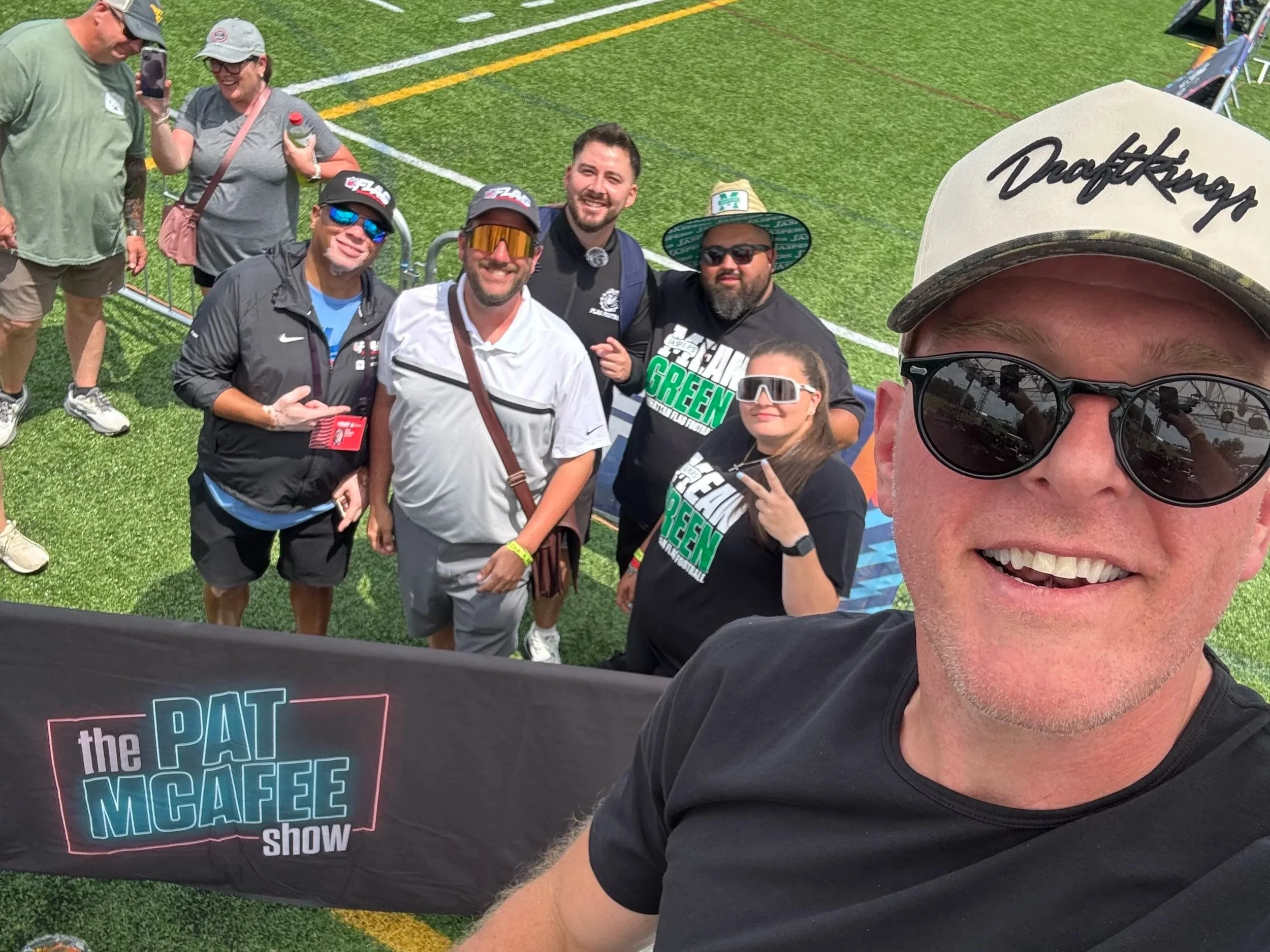A group of people, including a man in sunglasses and a white shirt and others wearing hats and sports shirts, gathered on a sports field for a selfie with a large sign that reads 'the PAT MCAFEE show.'