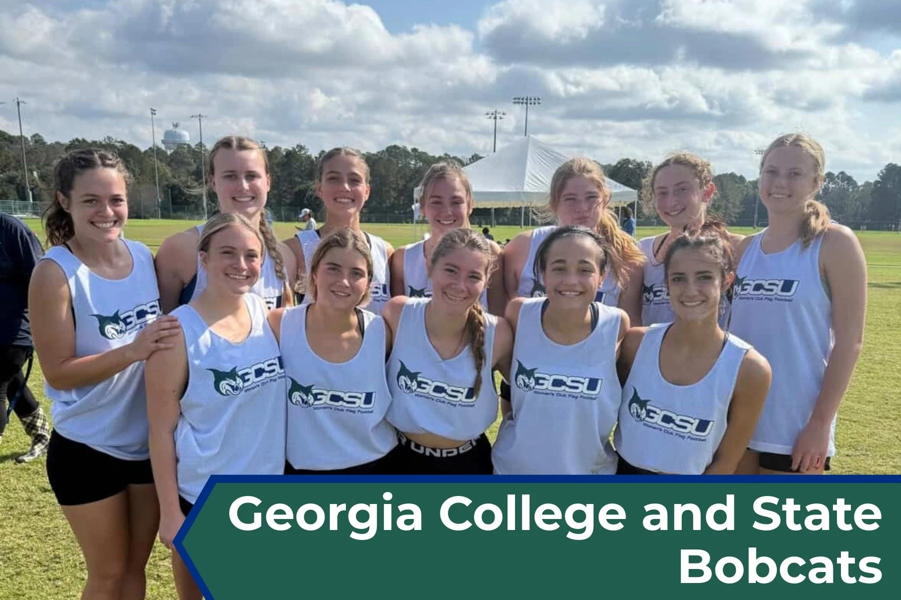 A group of 13 young women in athletic uniforms posing on a sports field with a tent and trees in the background. They are members of the Georgia College and State University Bobcats cross-country team.