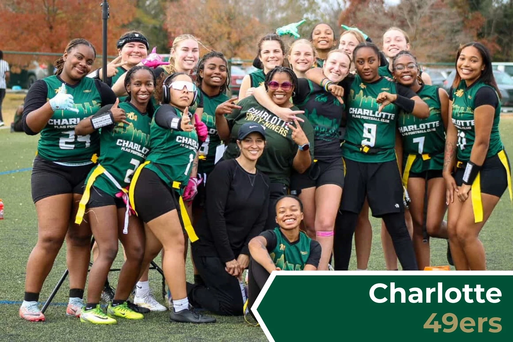 A group of female football players and their coach posing together on a football field, wearing green and black uniforms, celebrating after a game with some smiling, making peace signs and thumbs up gestures, with fall trees in the background. The bo