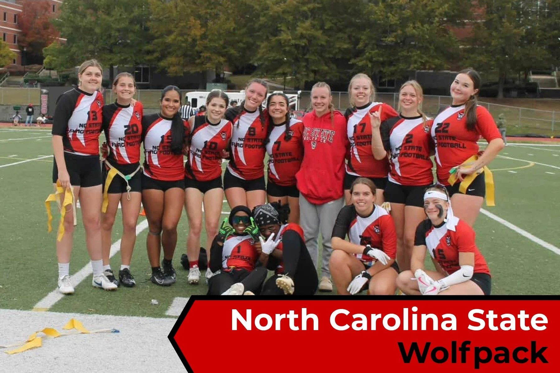 A group of female football players in red, black, and white uniforms with 'North Carolina State Football' written on them, posing together on a football field with a coach in the middle and a player on the ground in front. The background shows trees 