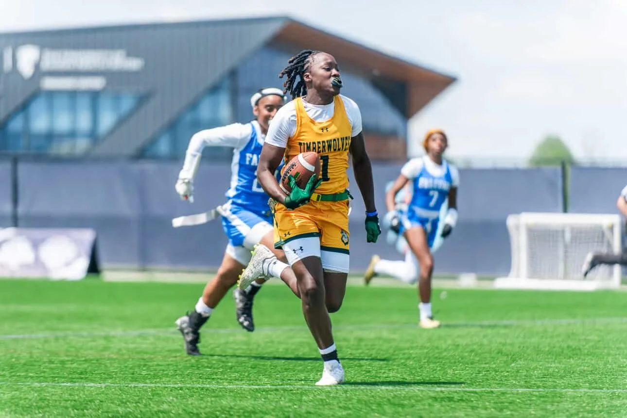 Women's College flag football player for Florida Gateway College in yellow uniform holding a football running on the field, with two other players in blue uniforms in the background.