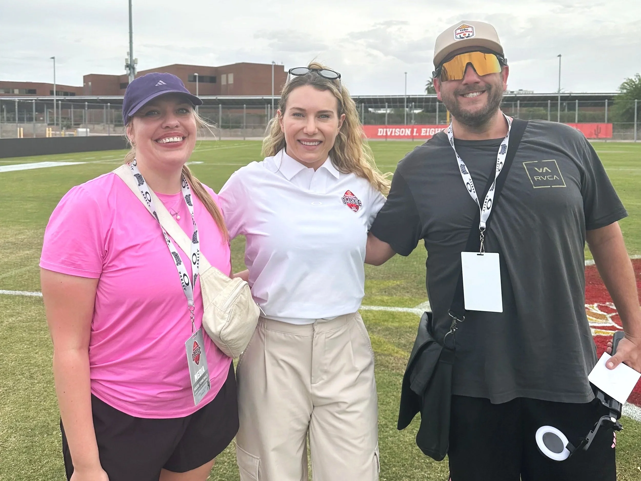 Three smiling people standing on a sports field with a chain-link fence and building in the background. The person on the left is wearing a purple hat and pink shirt, the person in the middle is in a white polo shirt with a logo, and the person on the right is wearing a beige cap, large reflective sunglasses, and a black t-shirt with a logo.
