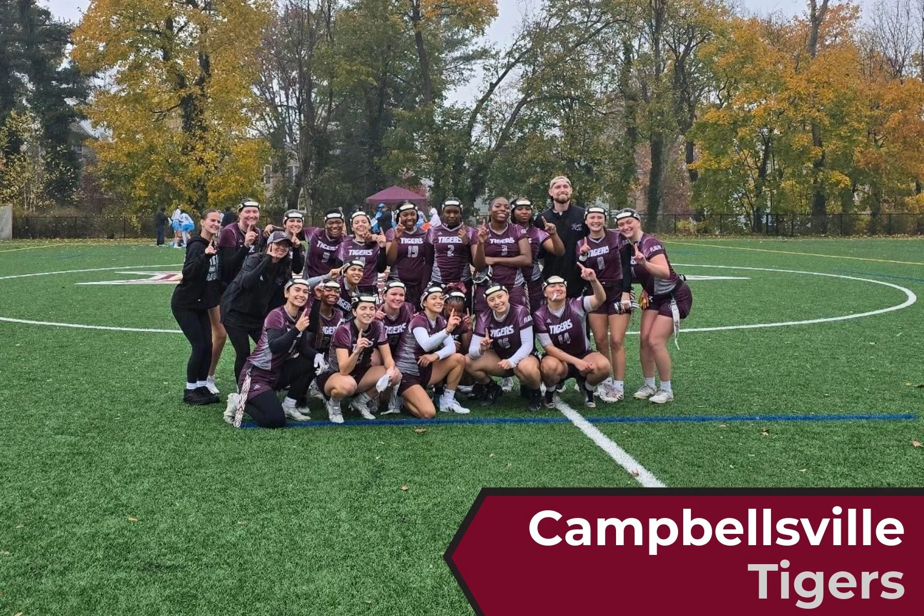 A group of female lacrosse players from the Campbellsiville Tigers team posing together with their coach on a soccer field. They are wearing maroon and gray uniforms, some are kneeling in front while others stand behind, and all are smiling and makin