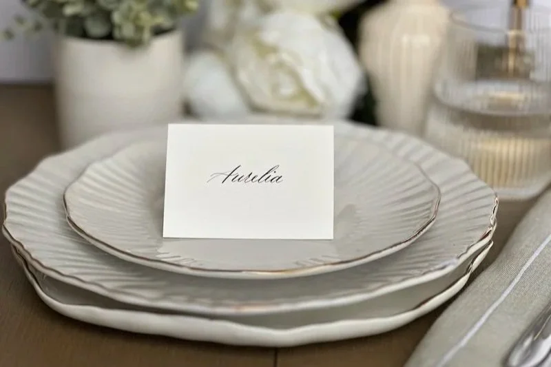 Elegant wedding place setting with a cream color place card with the name 'Aurelia', and decorative items in the background.