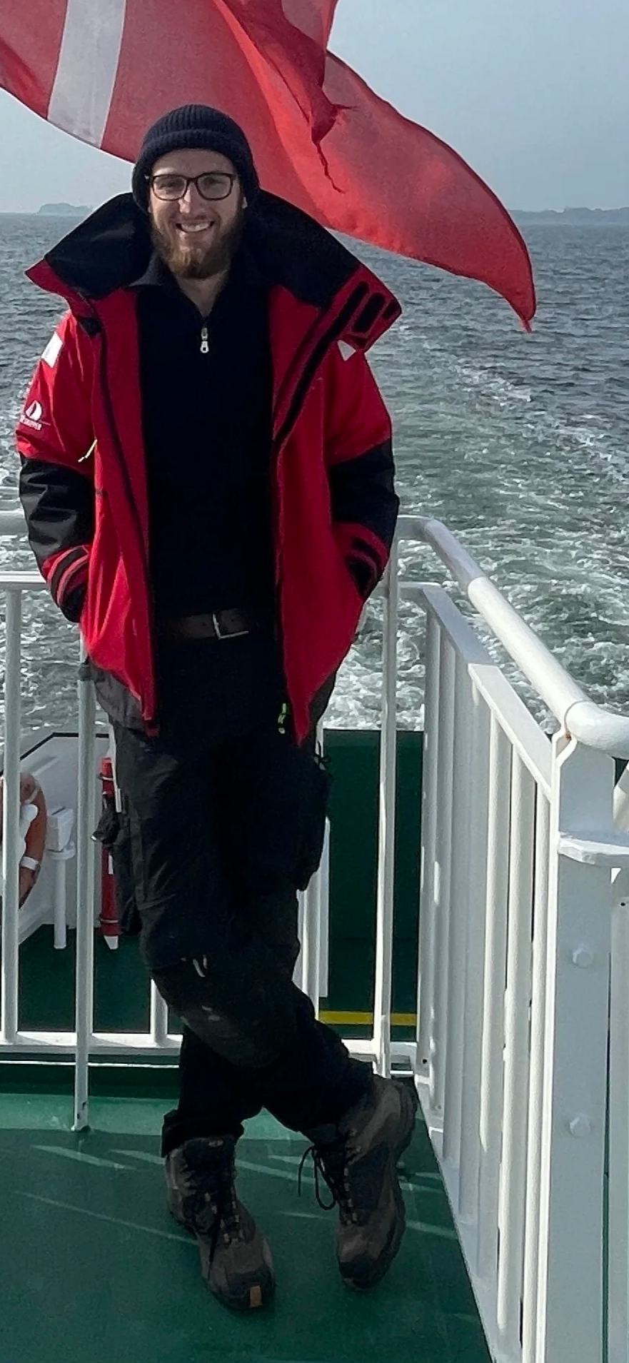 Mann in wetterfester Kleidung steht auf einem Boot, im Hintergrund ist das Meer zu sehen, und im Wind wehte eine rote Fahne.