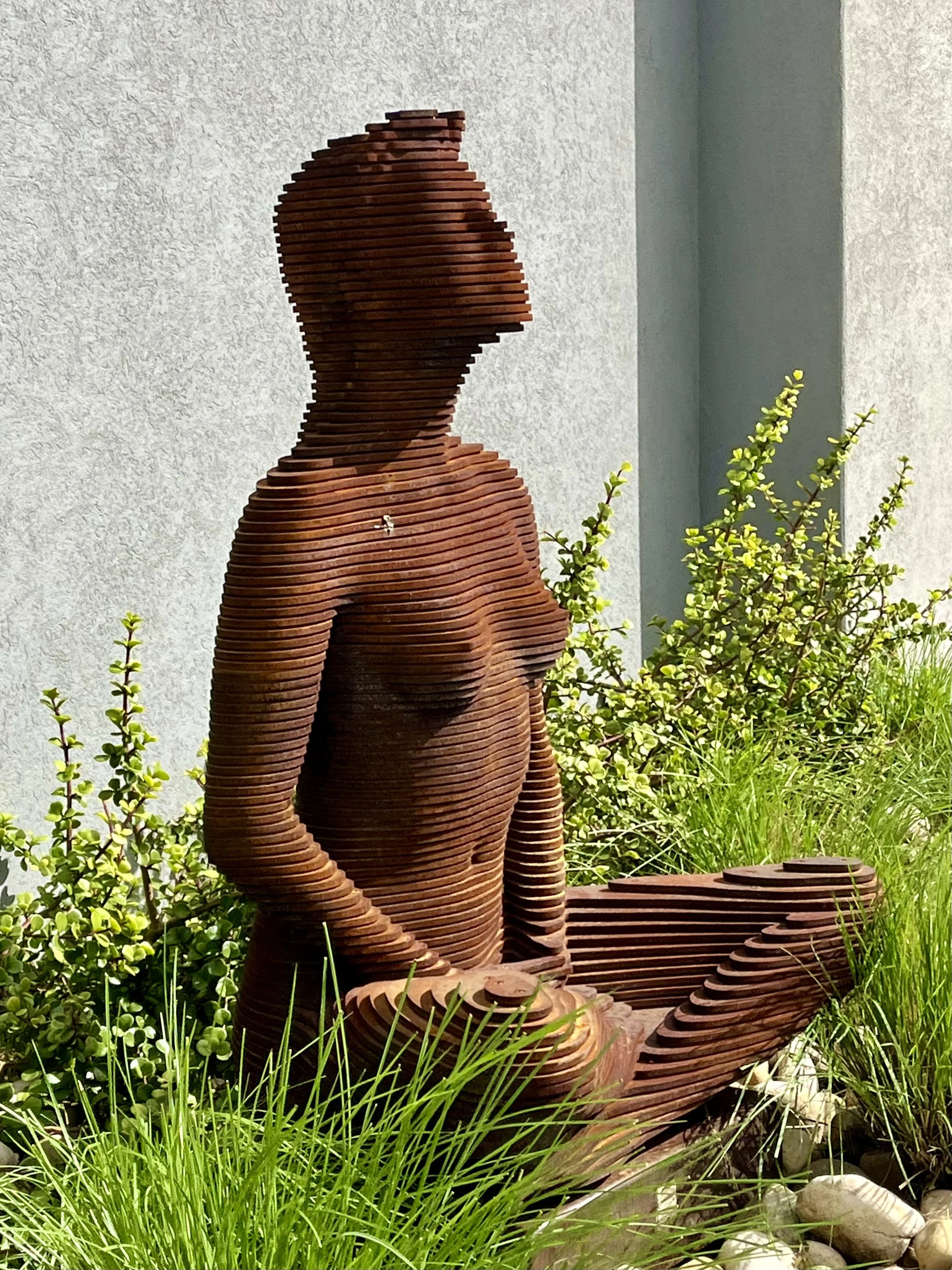 Layered metal sculpture of a seated human figure in a garden setting