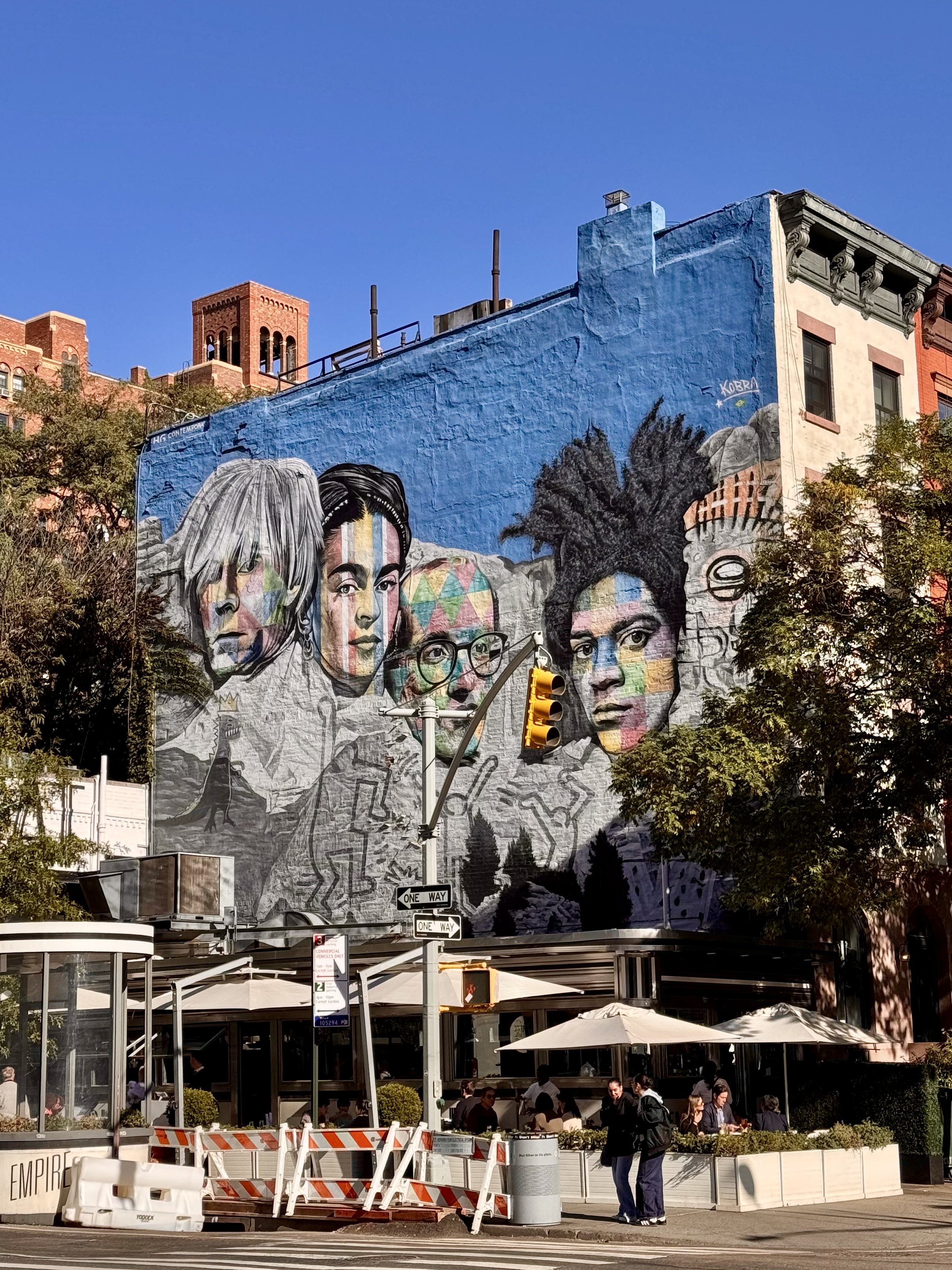 Colorful mural on a building depicting multiple faces in a busy urban area.