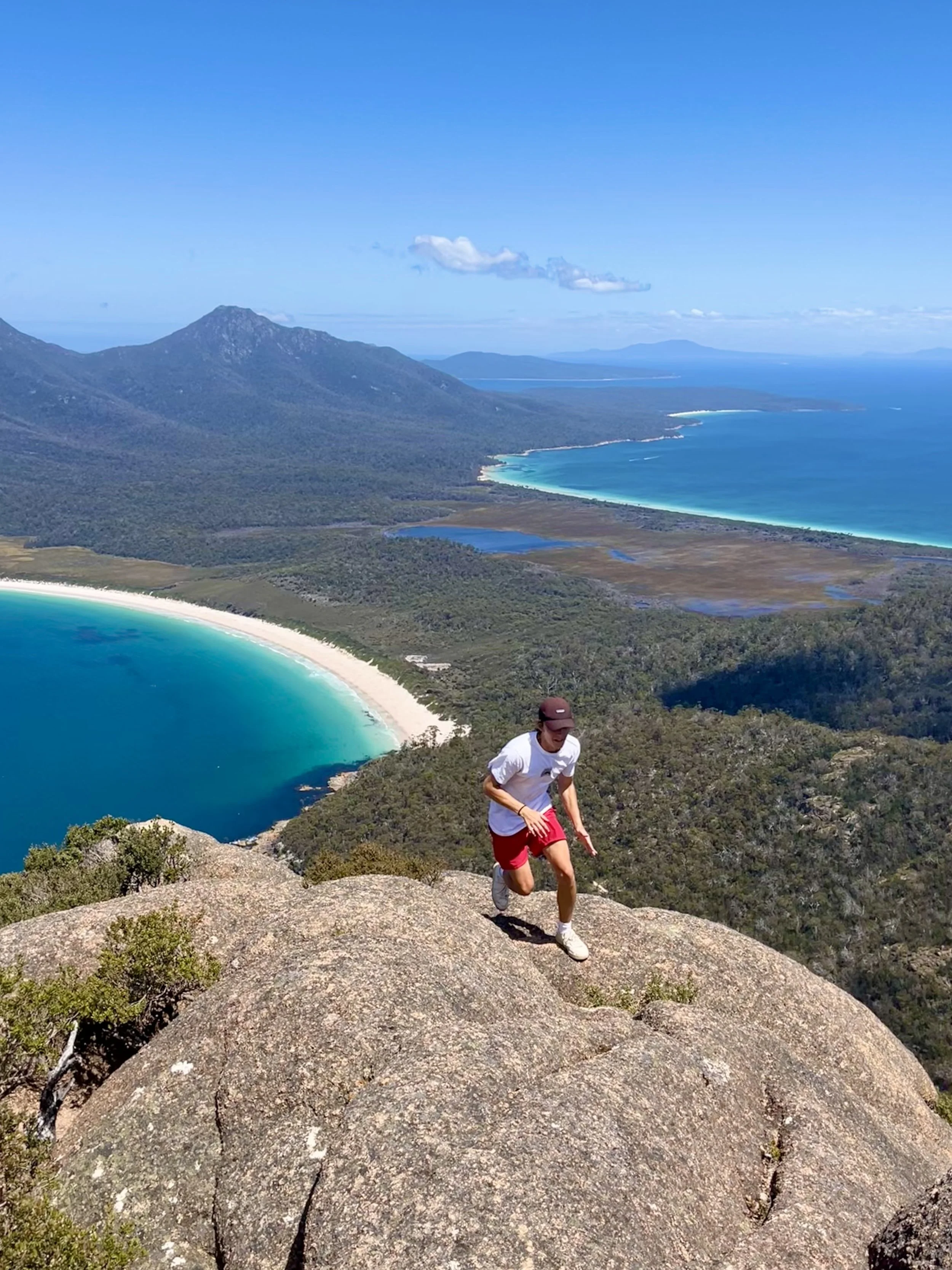 Person climbing rocky hill above scenic coastal landscape with blue ocean and sandy beach.