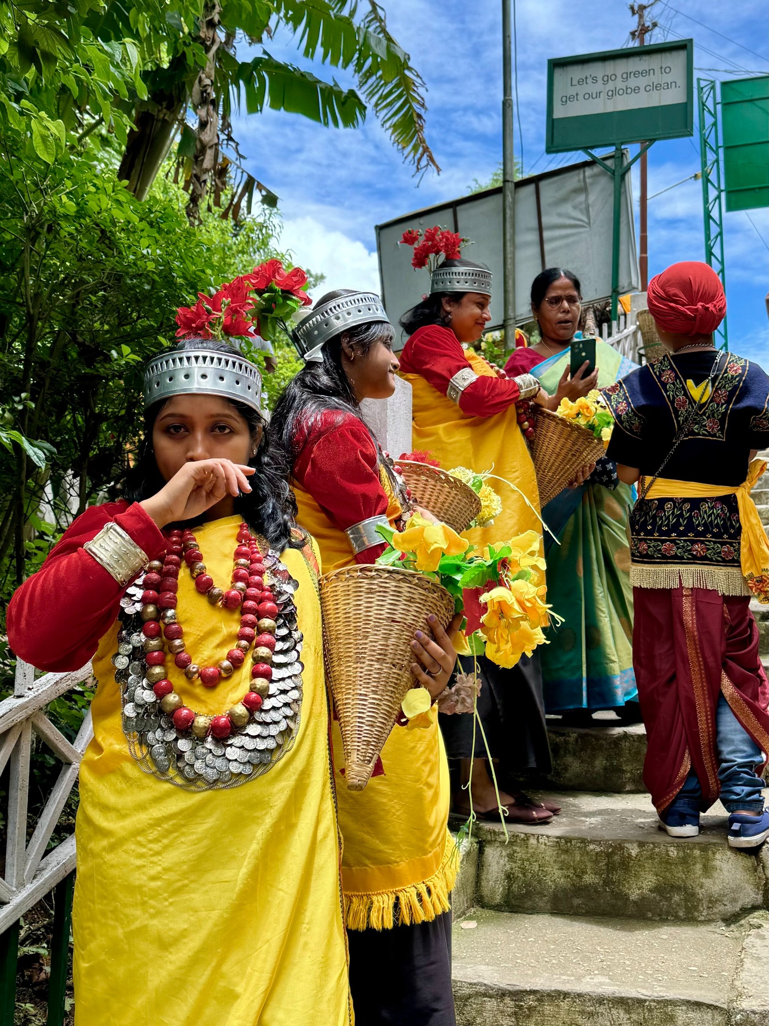 A group of people dressed in vibrant traditional attire, including yellow garments and ornate silver jewelry, standing on outdoor stairs. They are holding baskets with yellow flowers. The scene includes greenery and a sign that says 'Let's go green t