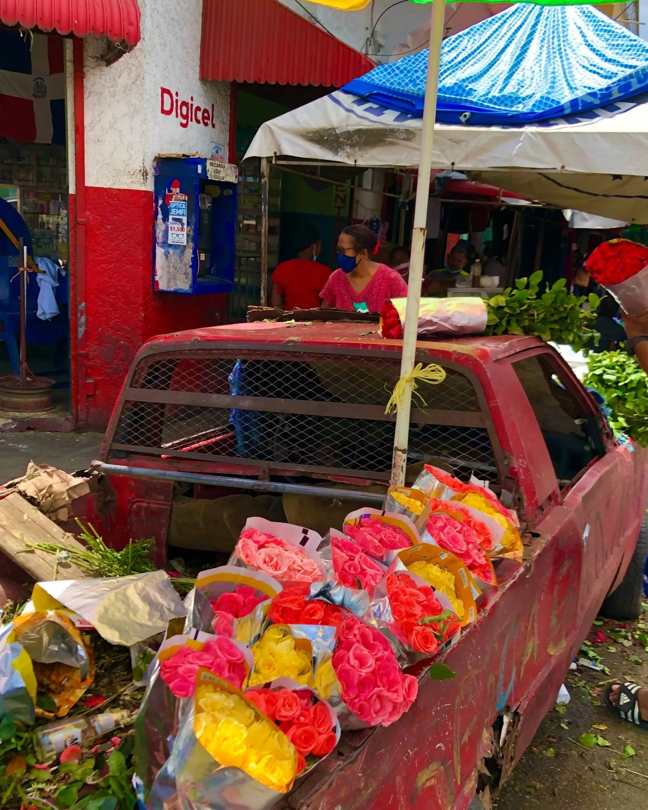 A red truck with colorful bouquets of roses, including pink, yellow, and red, parked outside a market stall under umbrellas. A person is holding a bouquet of red roses. The area is decorated with greenery and there is a sign for Digicel on the buildi