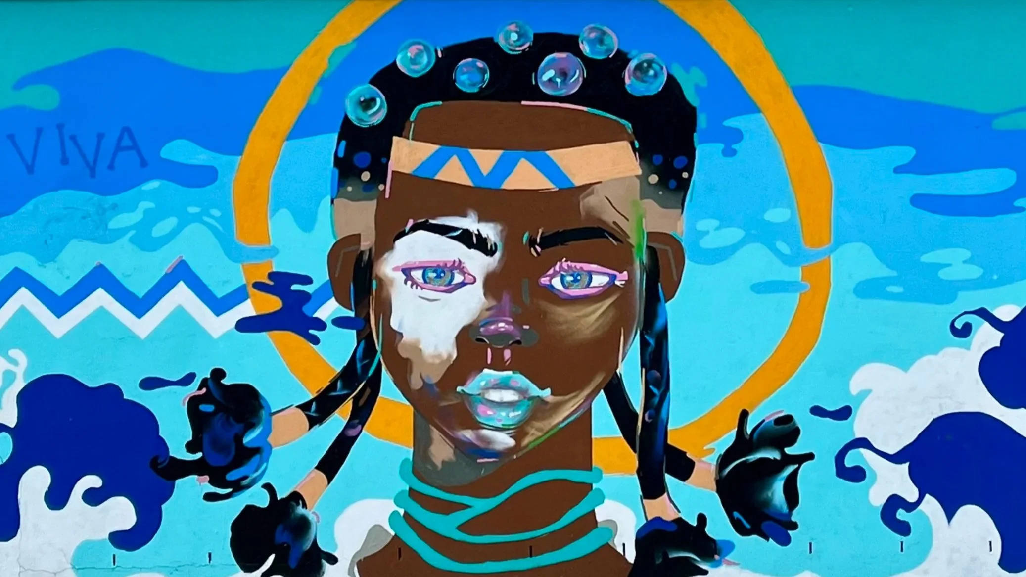 Street art mural featuring a stylized portrait of a person with colorful patterns and circular elements, set against a blue and orange background. Mountain landscape and cloudy sky visible in the distance.