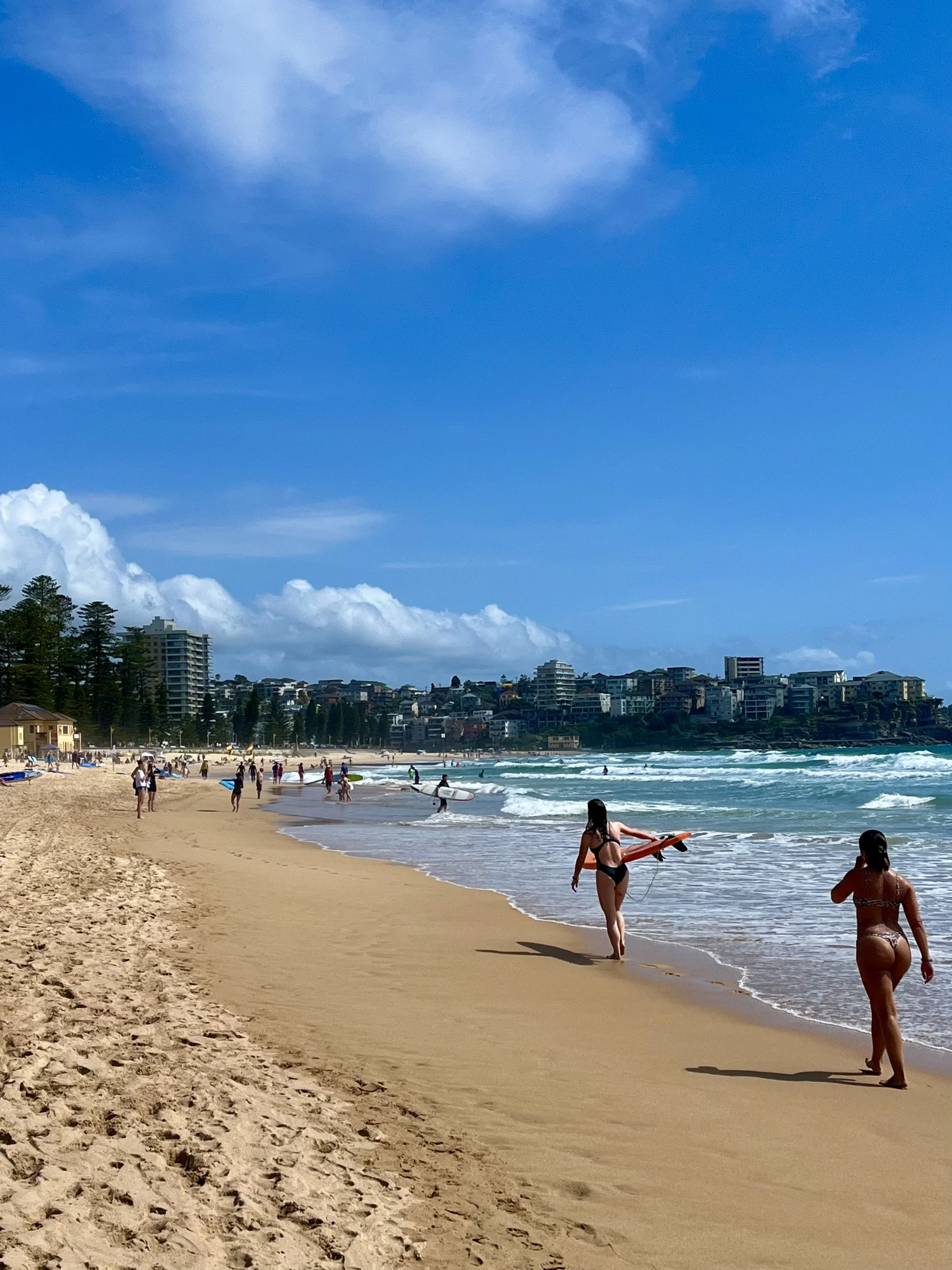 A sandy beach with people walking along the shore, one person carrying a surfboard. The ocean waves are visible, and there are buildings and trees in the background under a partly cloudy blue sky.