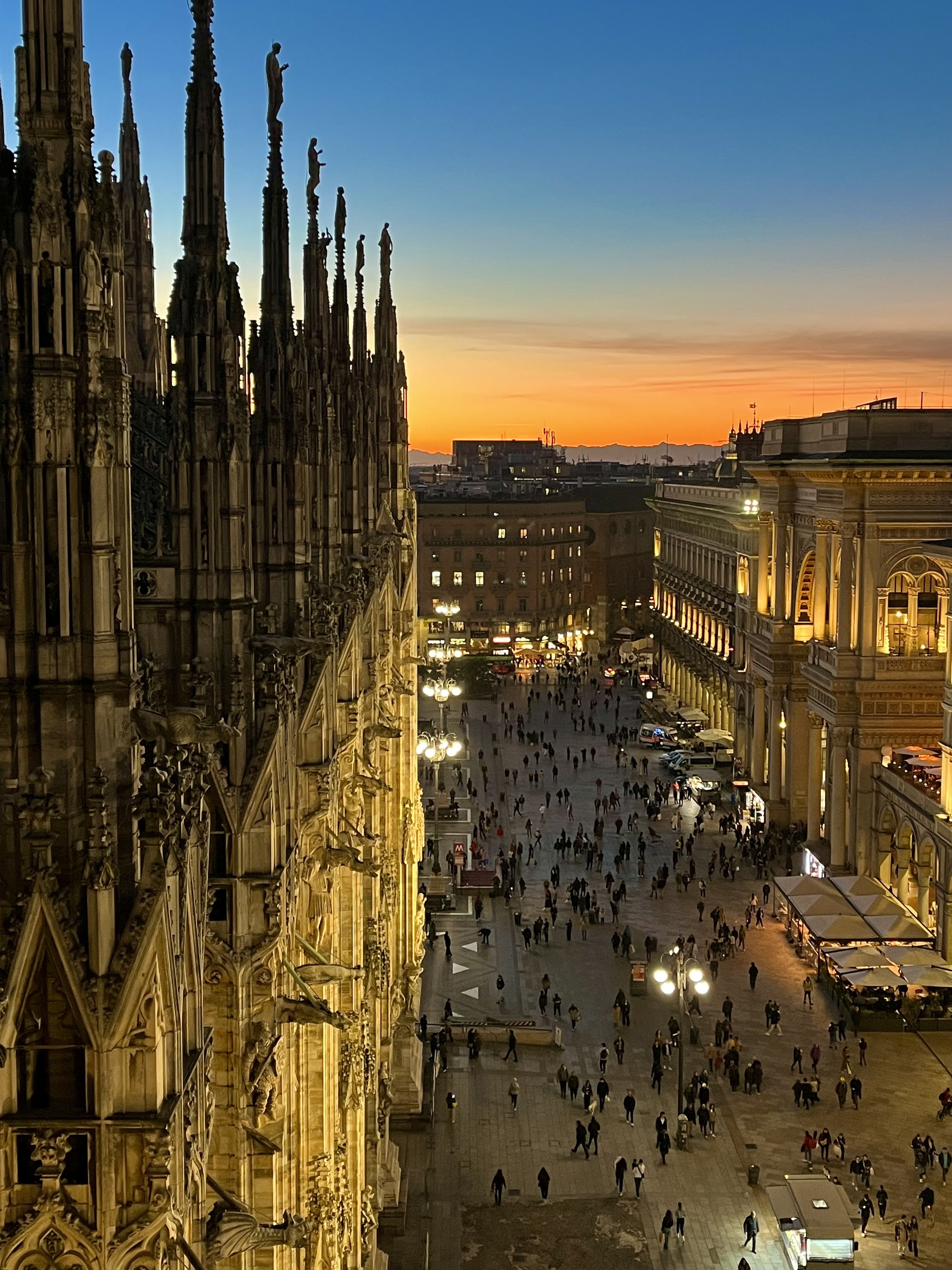 Evening view of Milan Cathedral with illuminated spires on the left and a busy city square below, against a sunset sky.