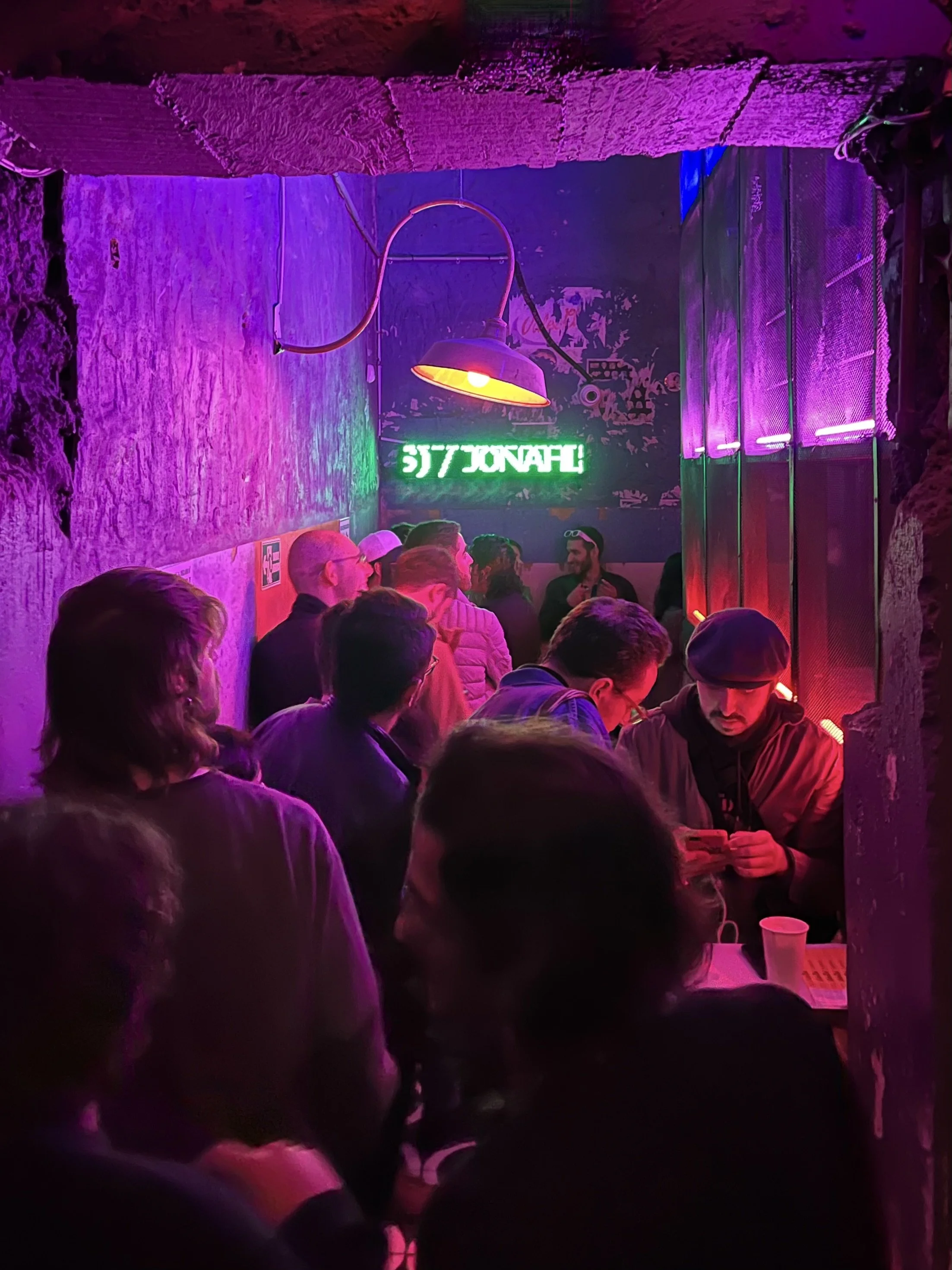 Crowded bar or club with purple lighting and a neon sign on the wall.