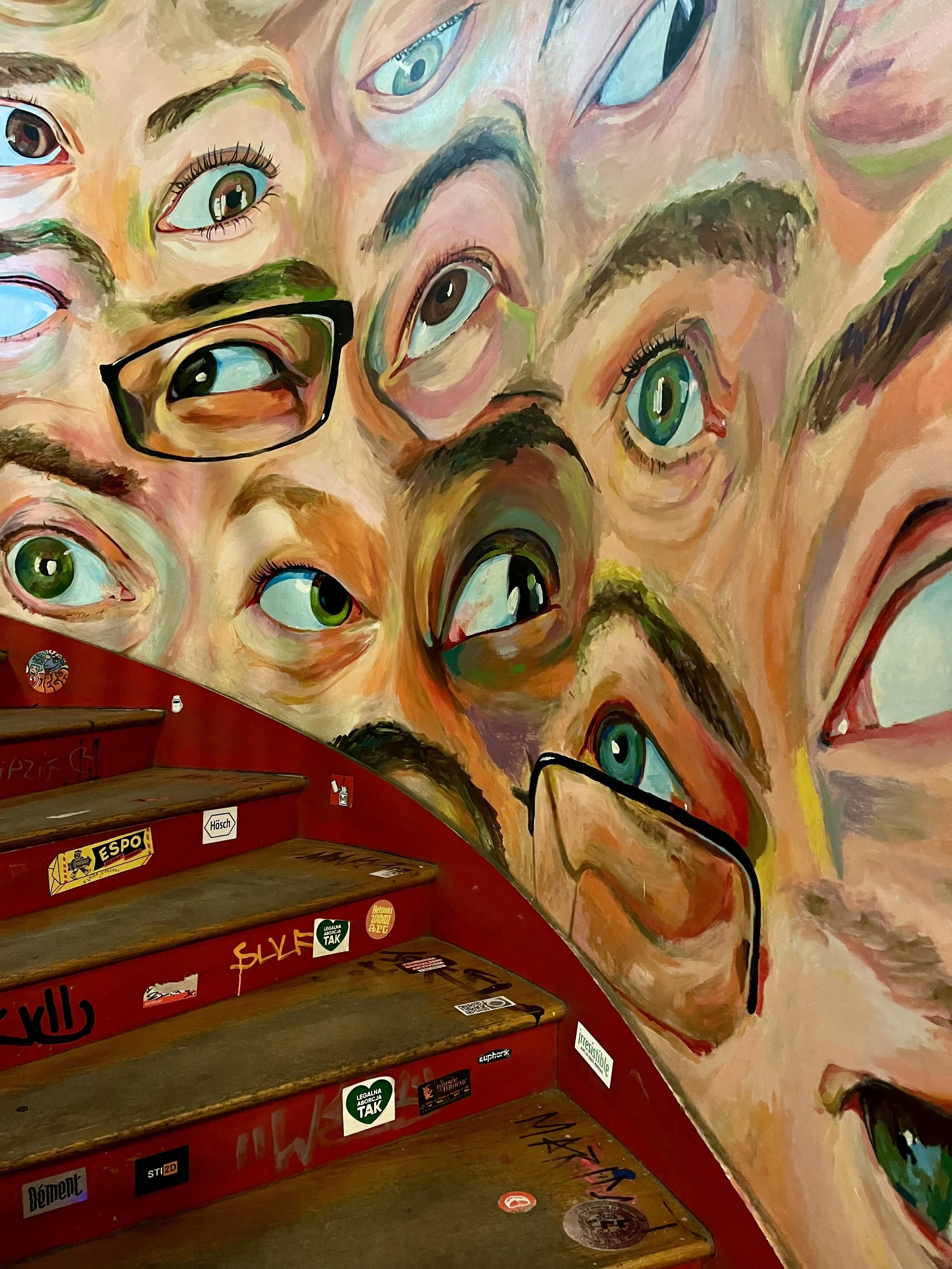 Colorful mural of multiple eyes on a wall beside red staircase with stickers.