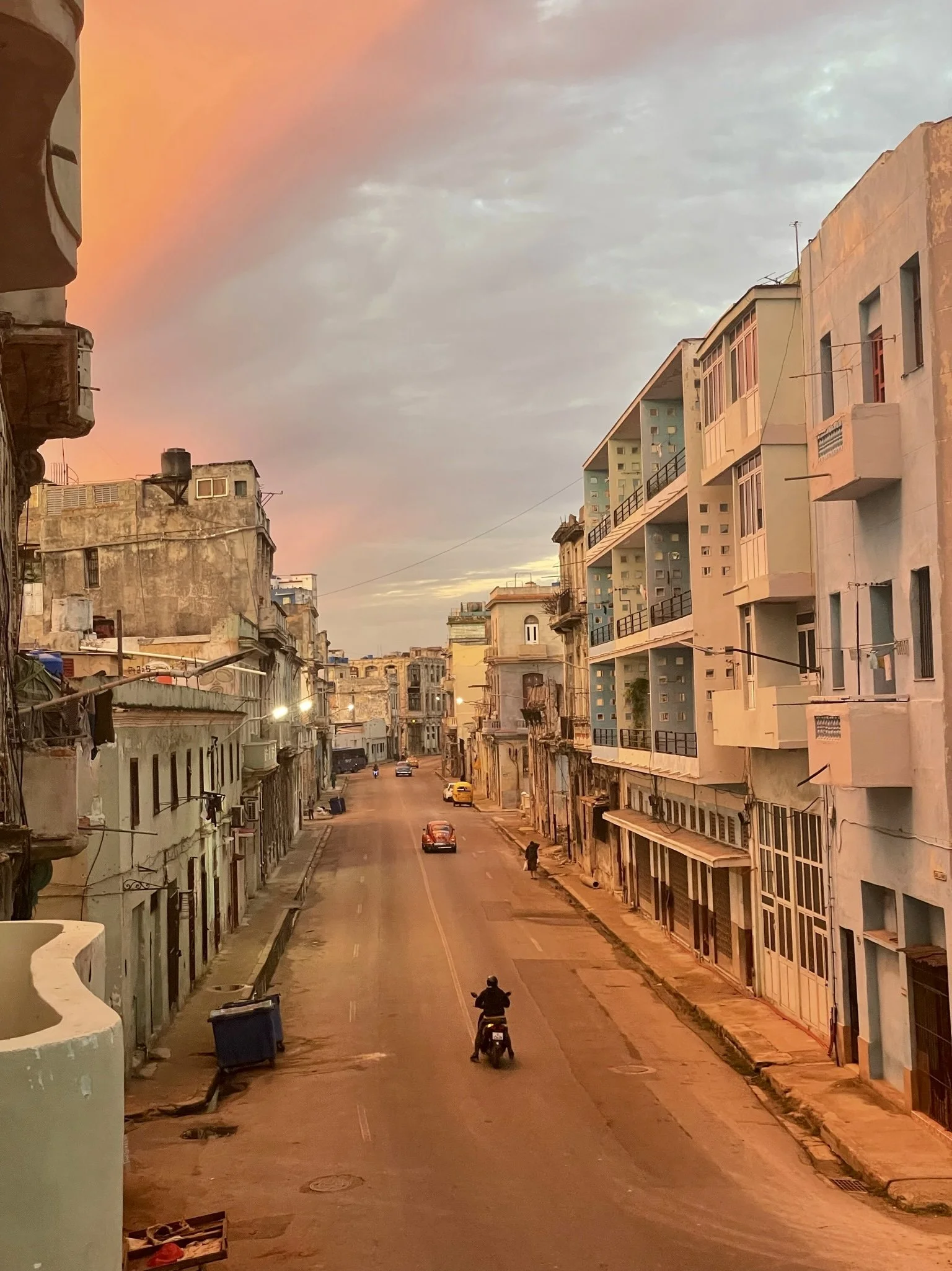 Street scene with old buildings and orange sky, featuring a person on a scooter and a vintage car.