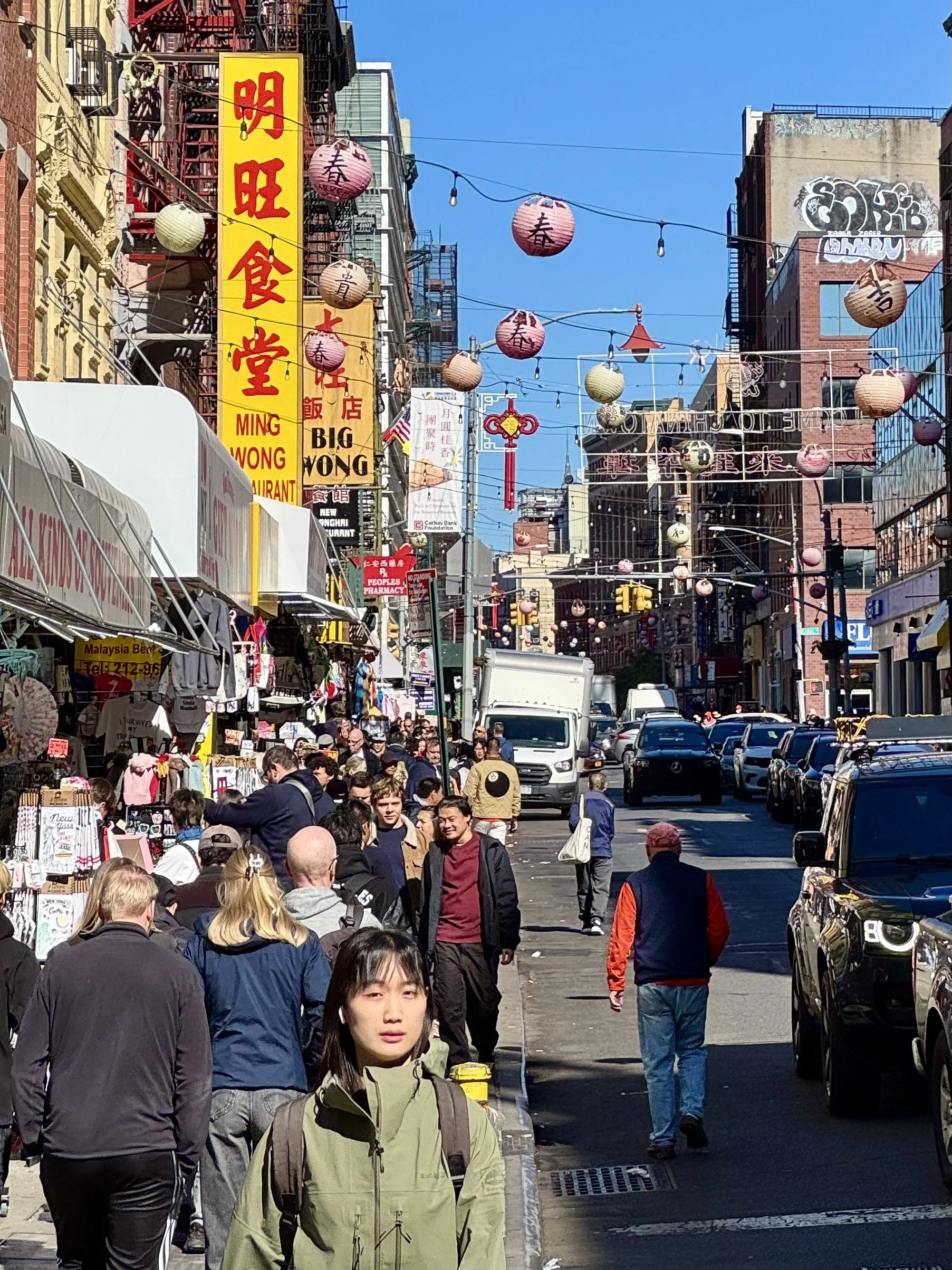 A busy street scene in Chinatown with colorful lanterns, diverse crowd, and storefronts featuring signs in Chinese and English.