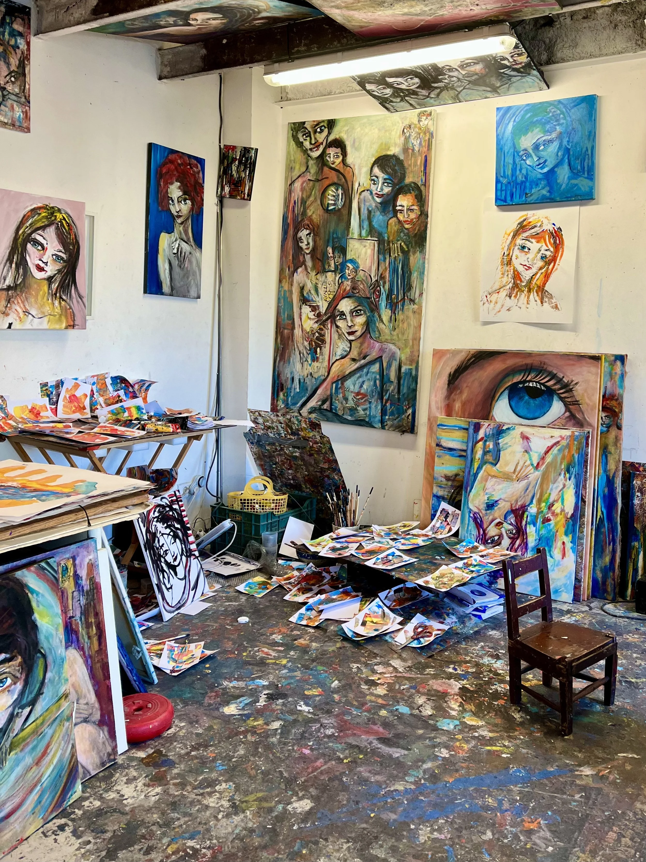 An artist's studio with colorful paintings on the walls and scattered on the floor. A wooden chair is in the center. The floor is covered in paint splatters. Various art materials and sketches are visible.