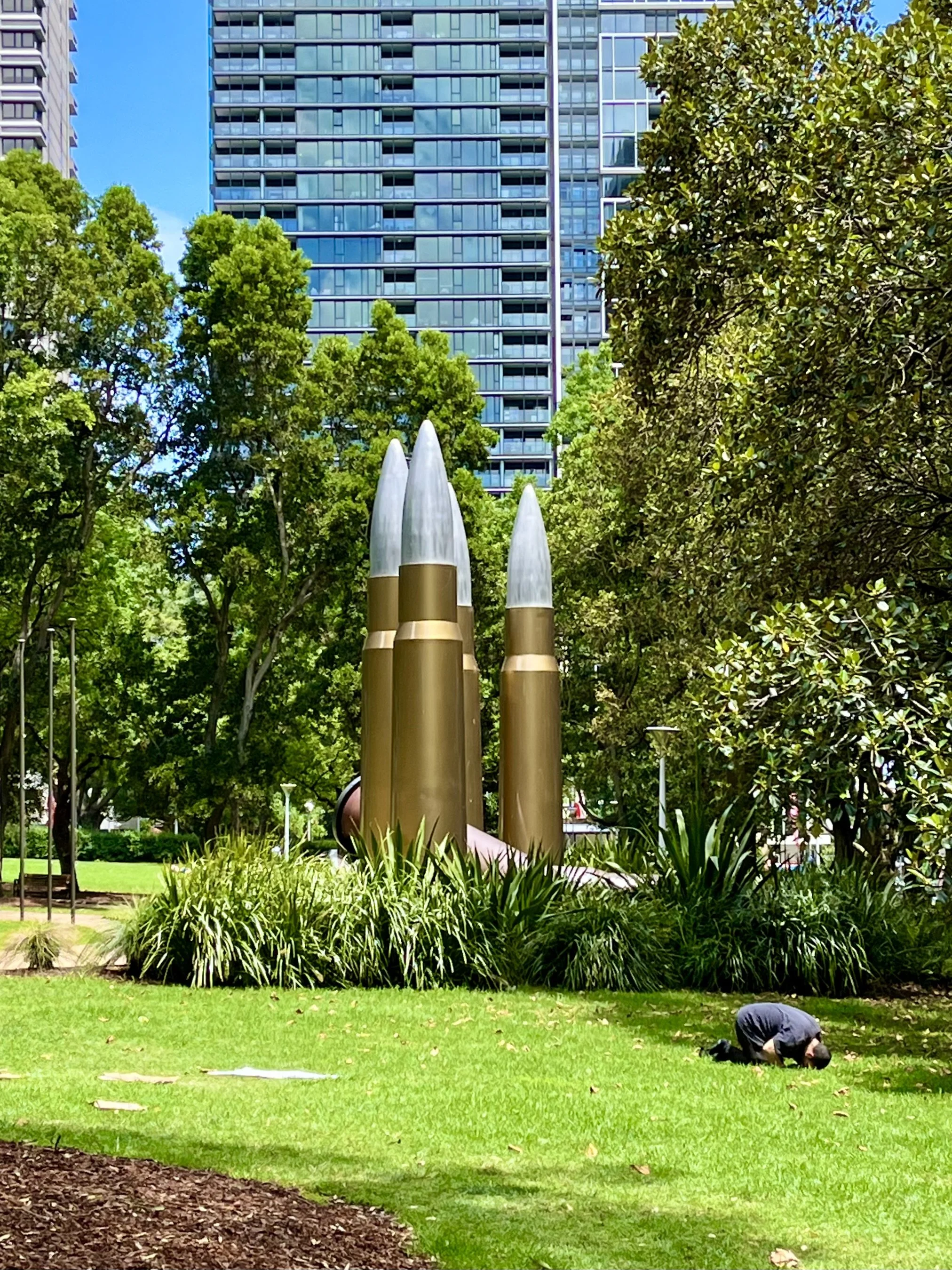 Park with large bullet sculptures, green grass, and city buildings in the background.