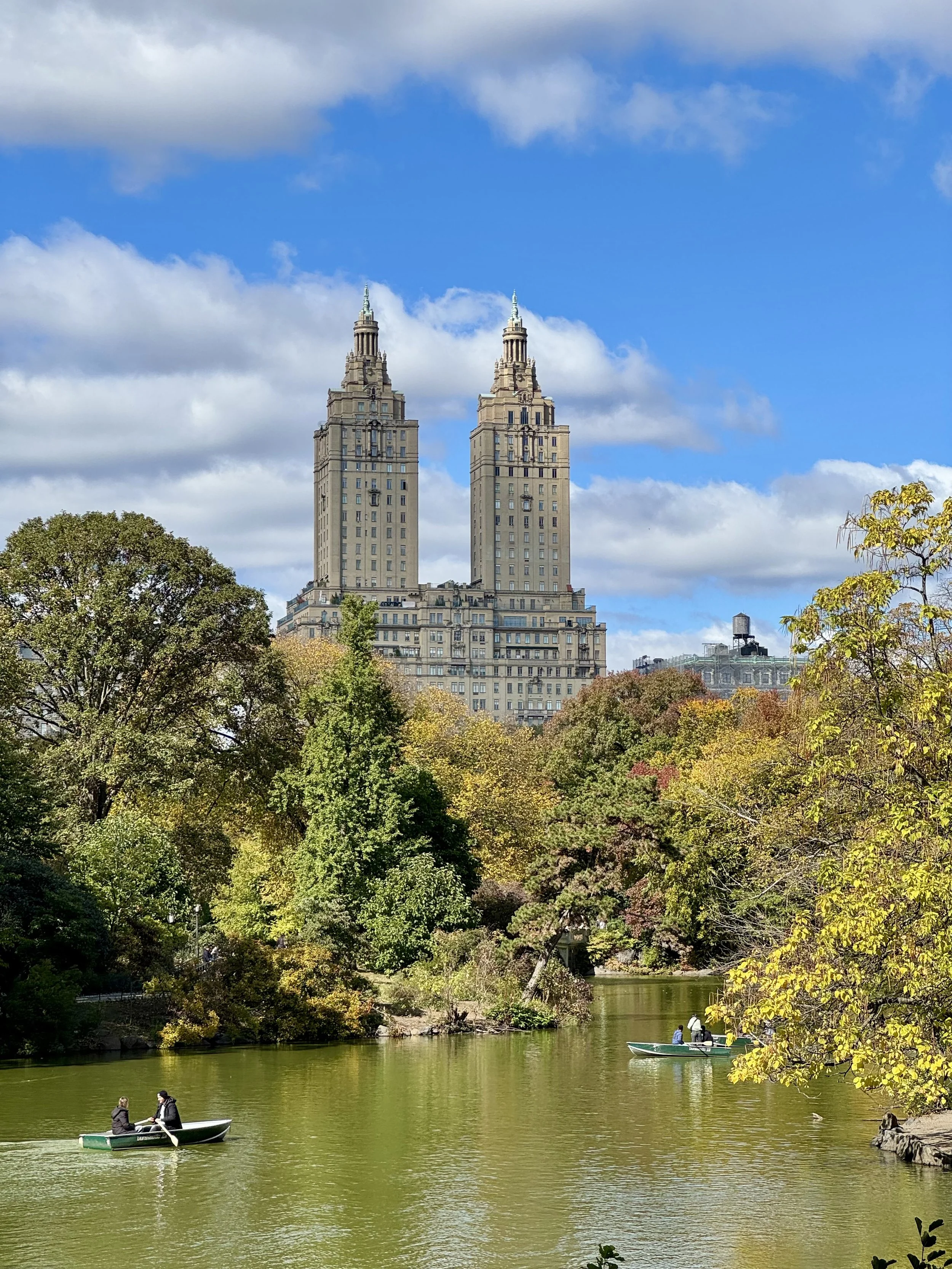 Twin-towered building with a pond in front, surrounded by trees, boats on the water, and a blue sky with clouds.