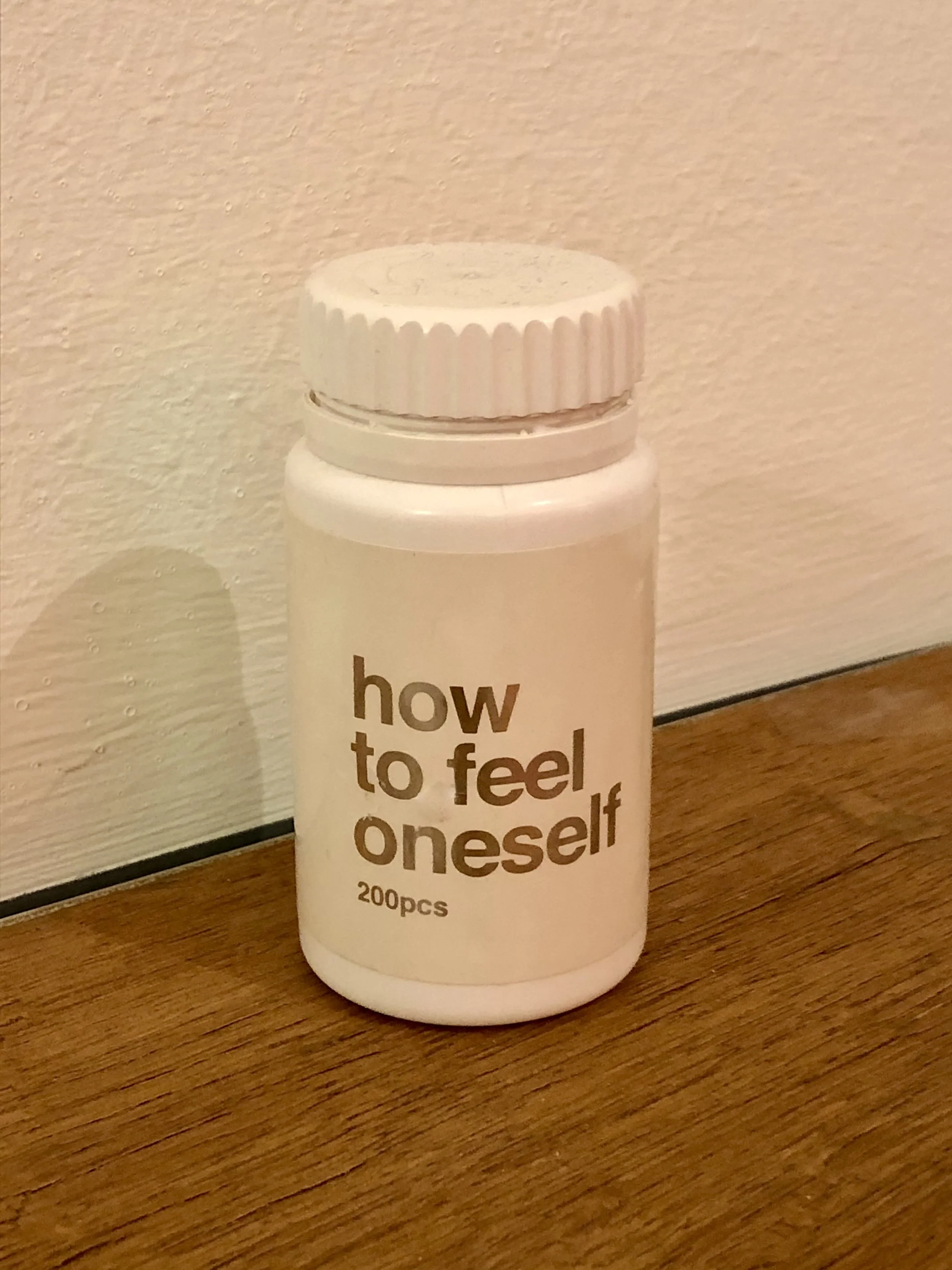 White pill bottle with label "how to feel oneself, 200pcs" placed on a wooden surface.