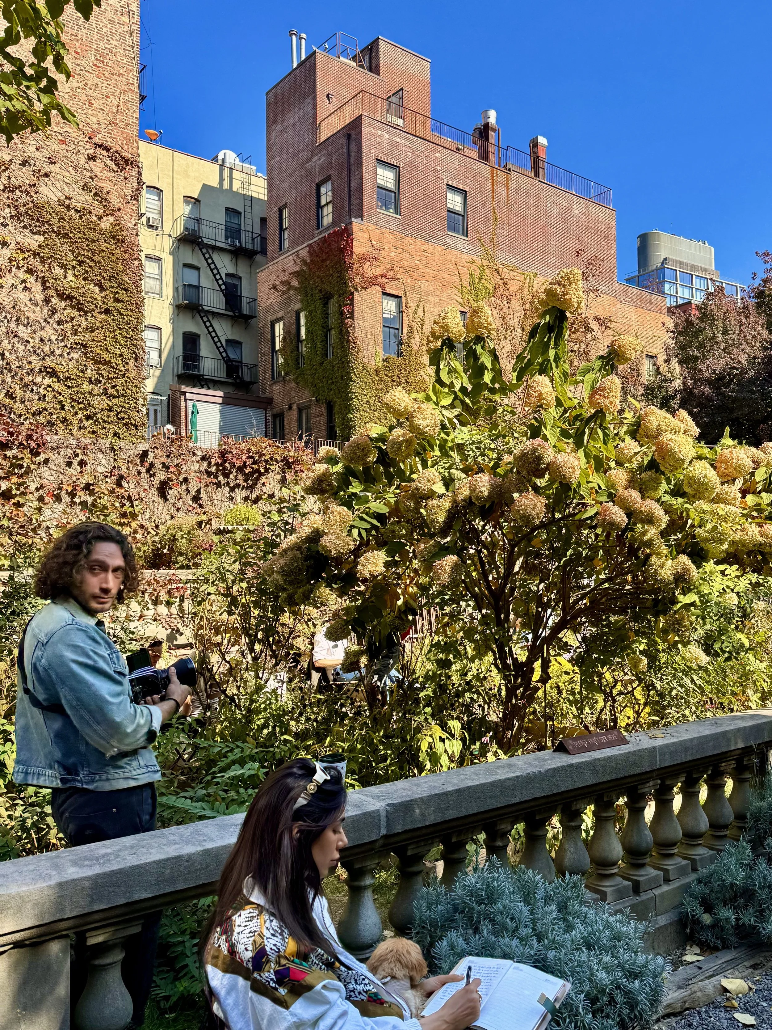 Urban garden with a person writing on a bench, another person holding a camera, and brick buildings in the background.