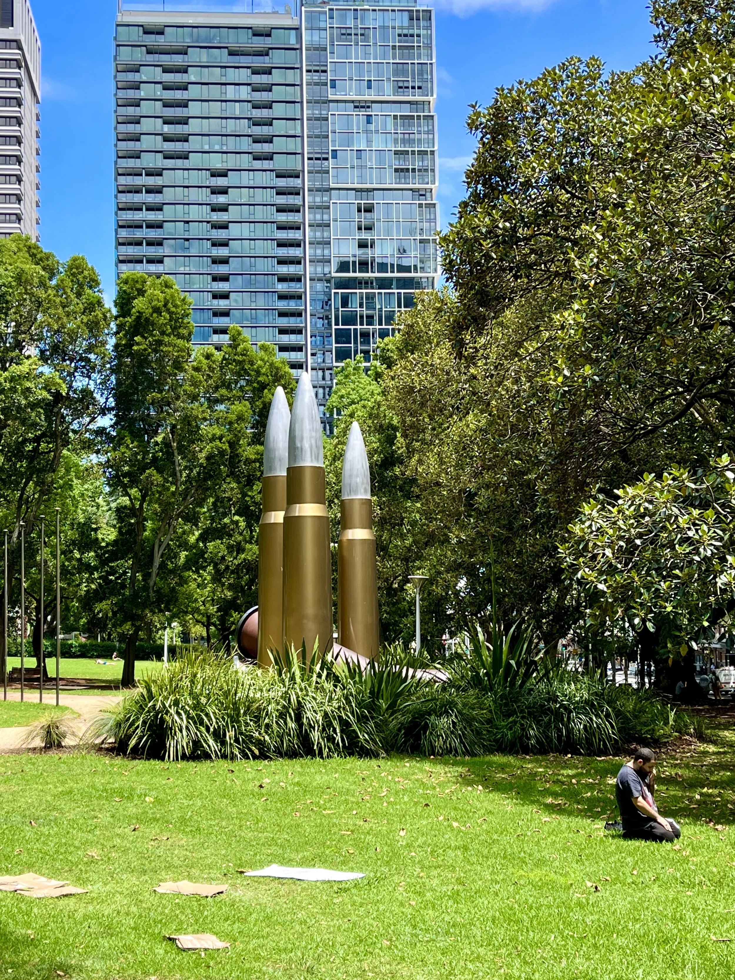 A park with tall green trees and modern high-rise buildings in the background. In the foreground, there are gold and silver metallic sculptures resembling bullets, surrounded by grass and bushes. A person is sitting on the grass to the right of the s