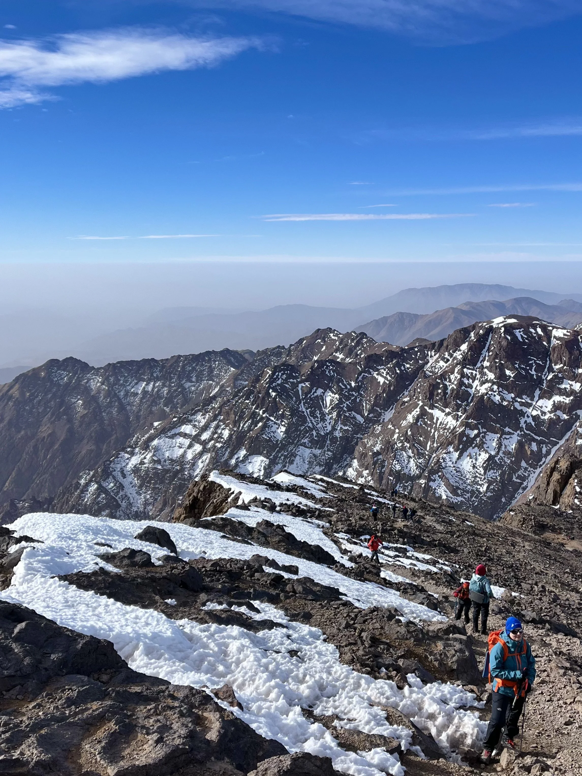 Hikers climbing a snowy mountain trail with rocky terrain under a clear blue sky.