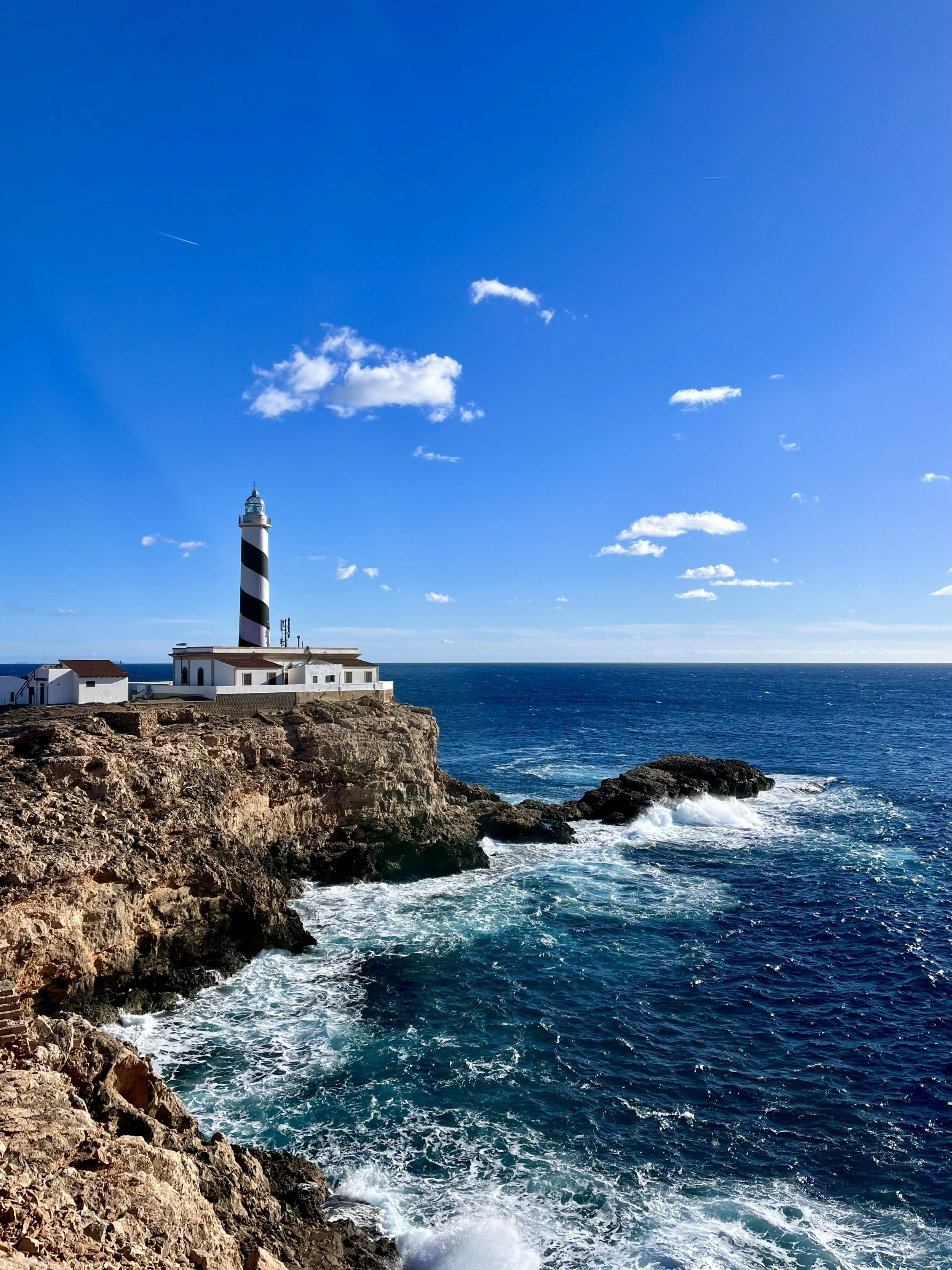 Lighthouse on rocky coastline with waves crashing, under a blue sky with a few clouds.