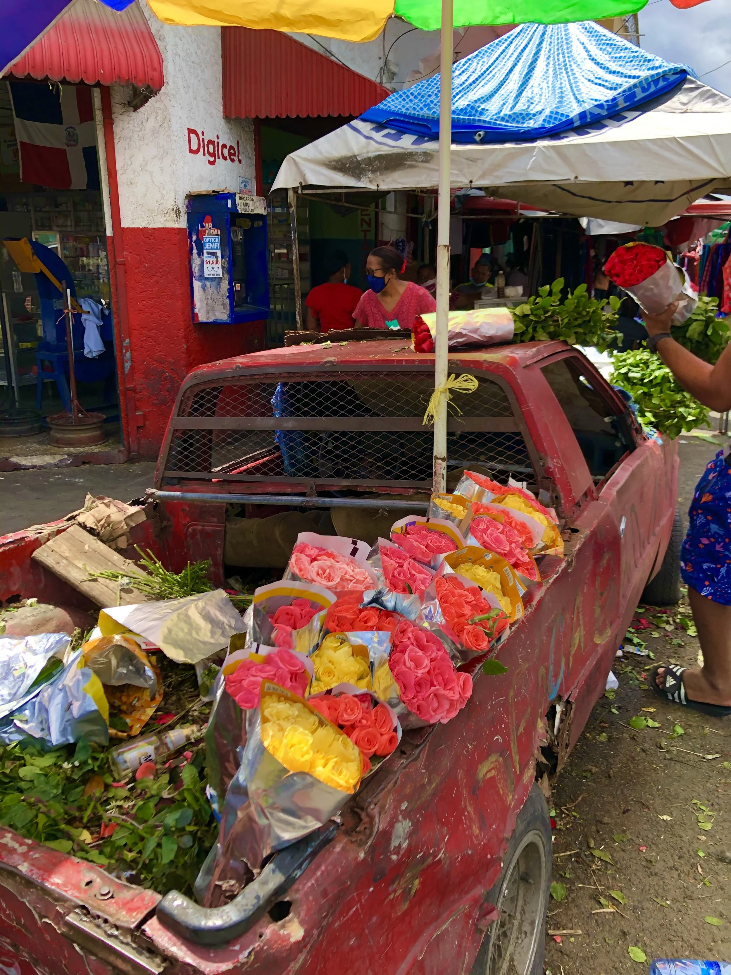 A red truck with colorful bouquets of roses, including pink, yellow, and red, parked outside a market stall under umbrellas. A person is holding a bouquet of red roses. The area is decorated with greenery and there is a sign for Digicel on the buildi