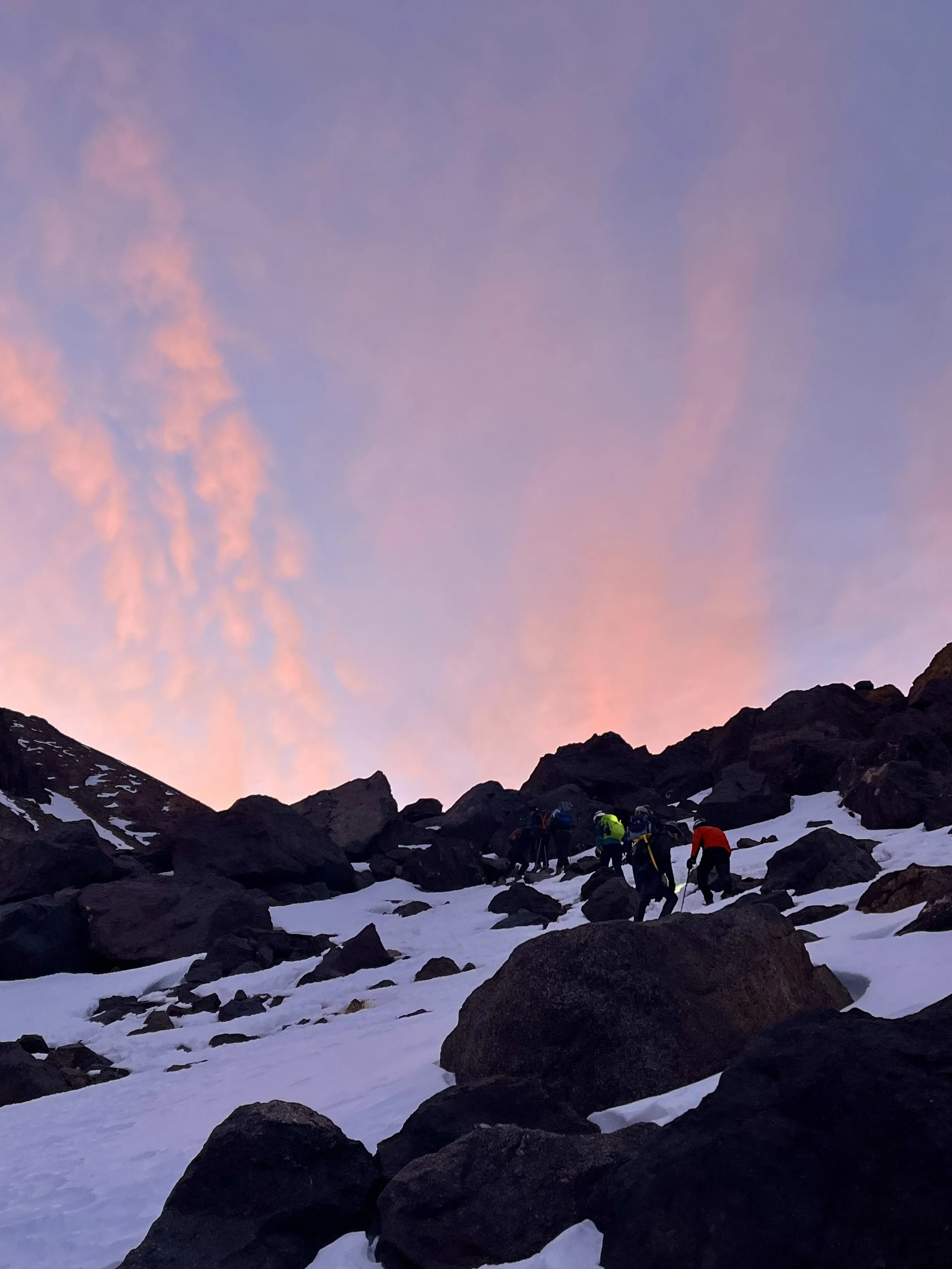 Group of climbers ascending a snow-covered mountain slope under a pink and blue sky at sunrise.