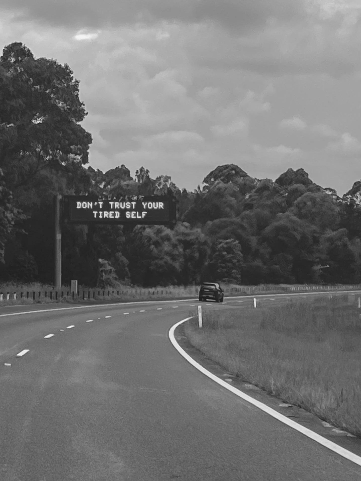 Road with a car driving under a sign displaying the message 'Don't trust your tired self' in a forested area.