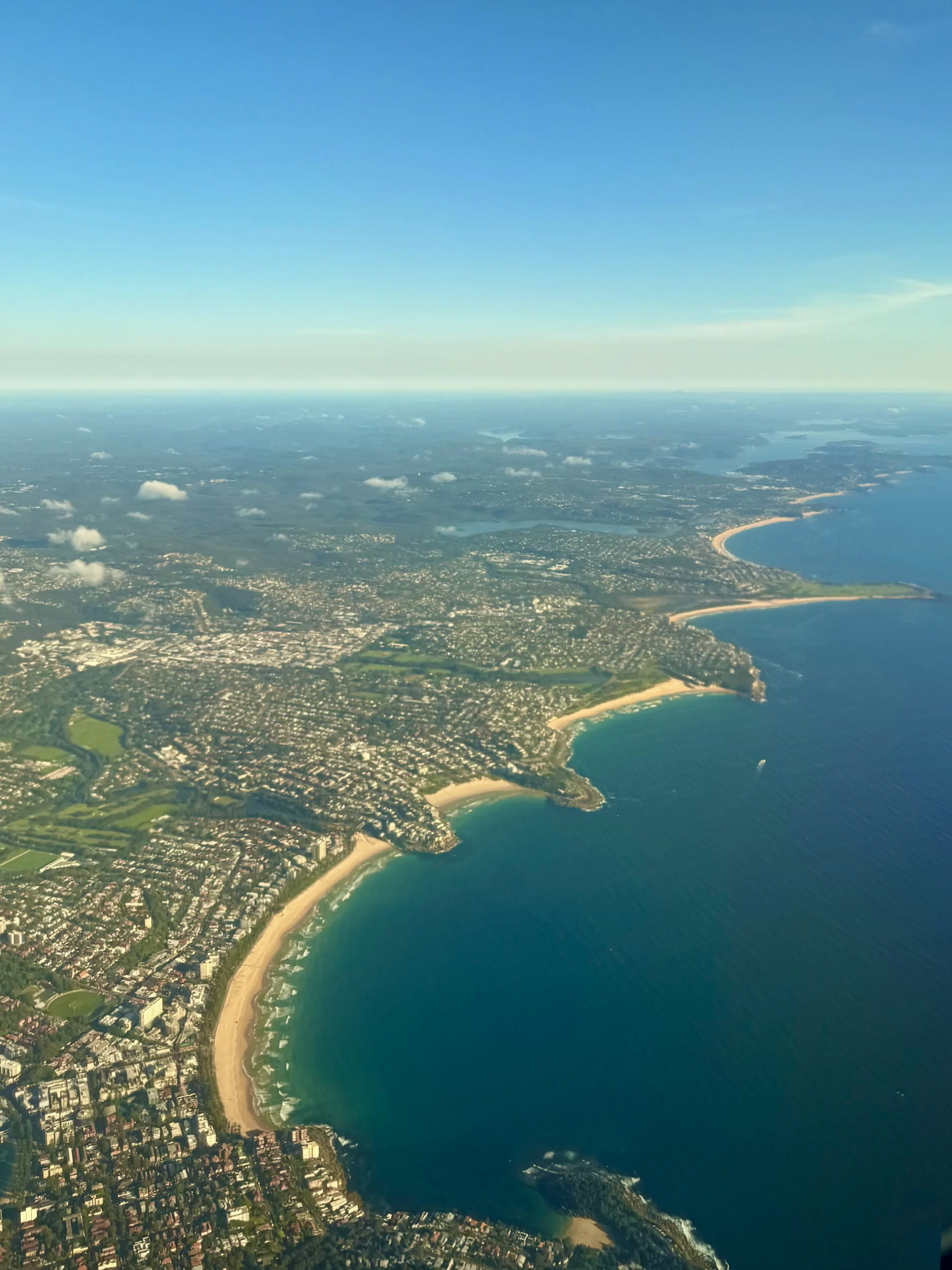 Aerial view of Australian coastal cityscape with multiple beaches and buildings, blue sea, and horizon under a clear sky.