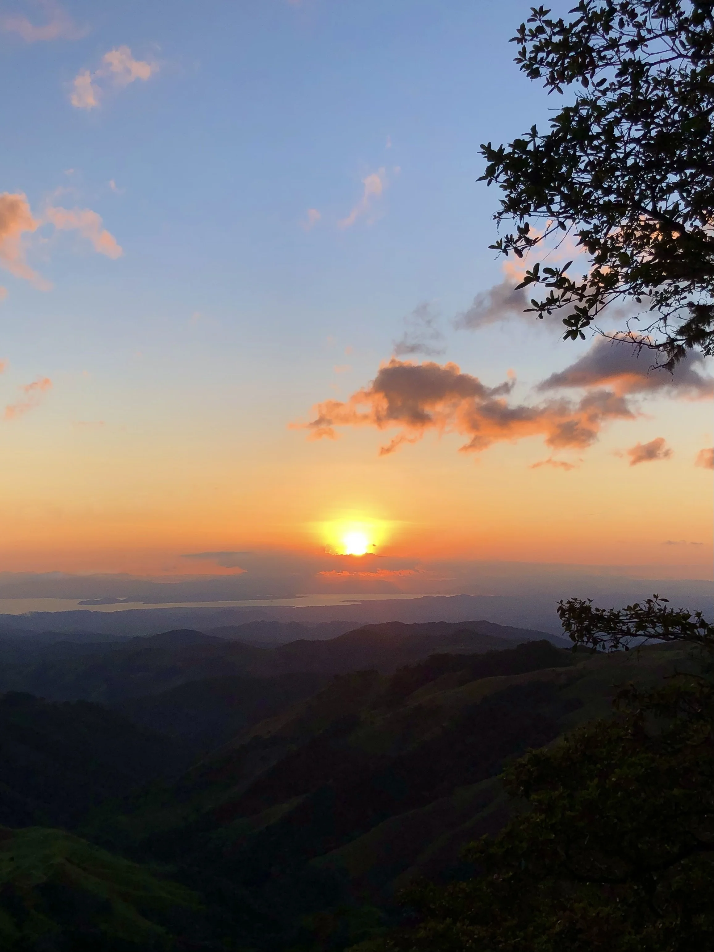 Sunset over a mountain landscape with a tree silhouette and scattered clouds in the sky.