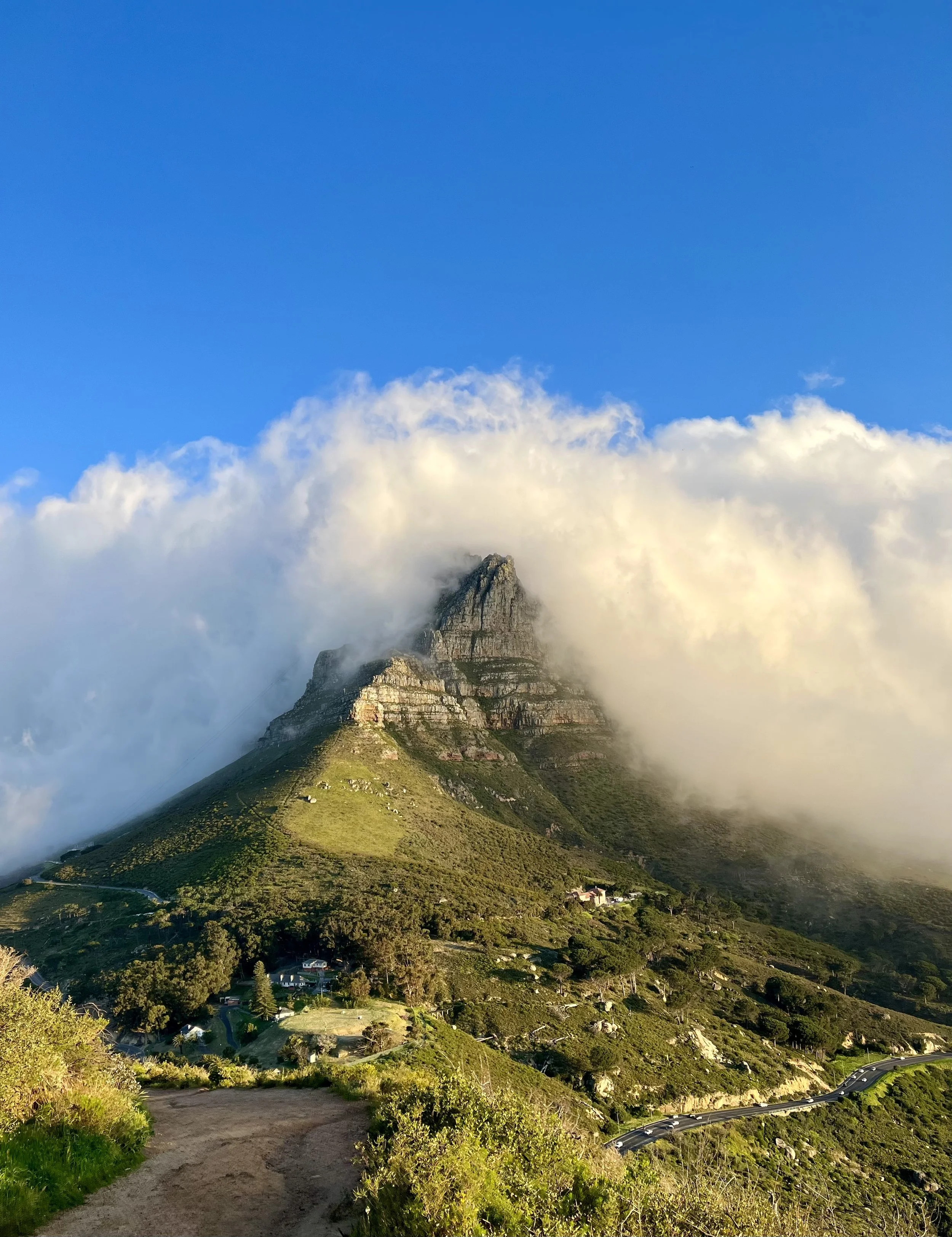 Mountain peak partially covered by clouds against a blue sky, with green vegetation on slopes and a winding path at the bottom.
