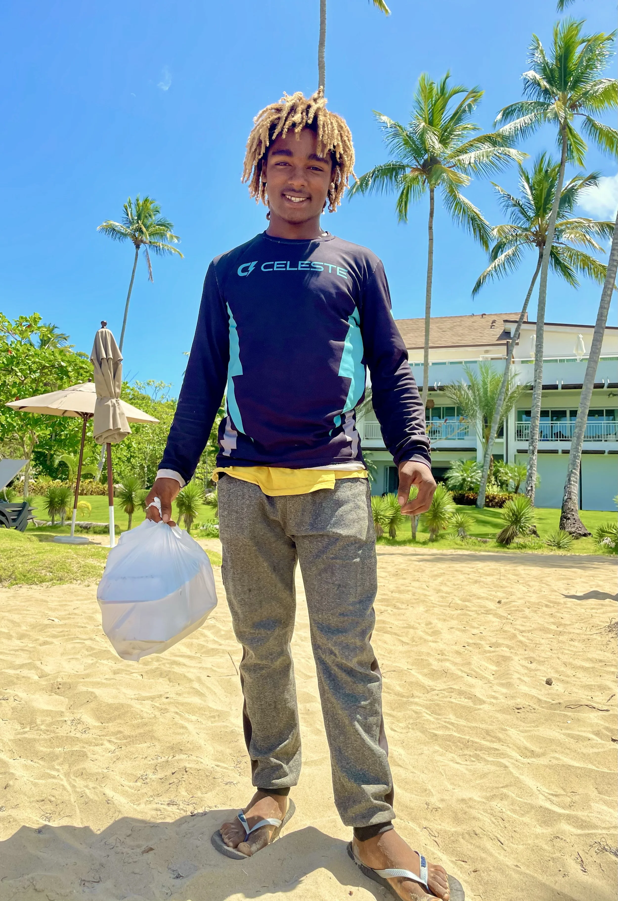 Person in a long-sleeve shirt standing on a sandy beach holding a plastic bag, surrounded by palm trees, under a clear blue sky.