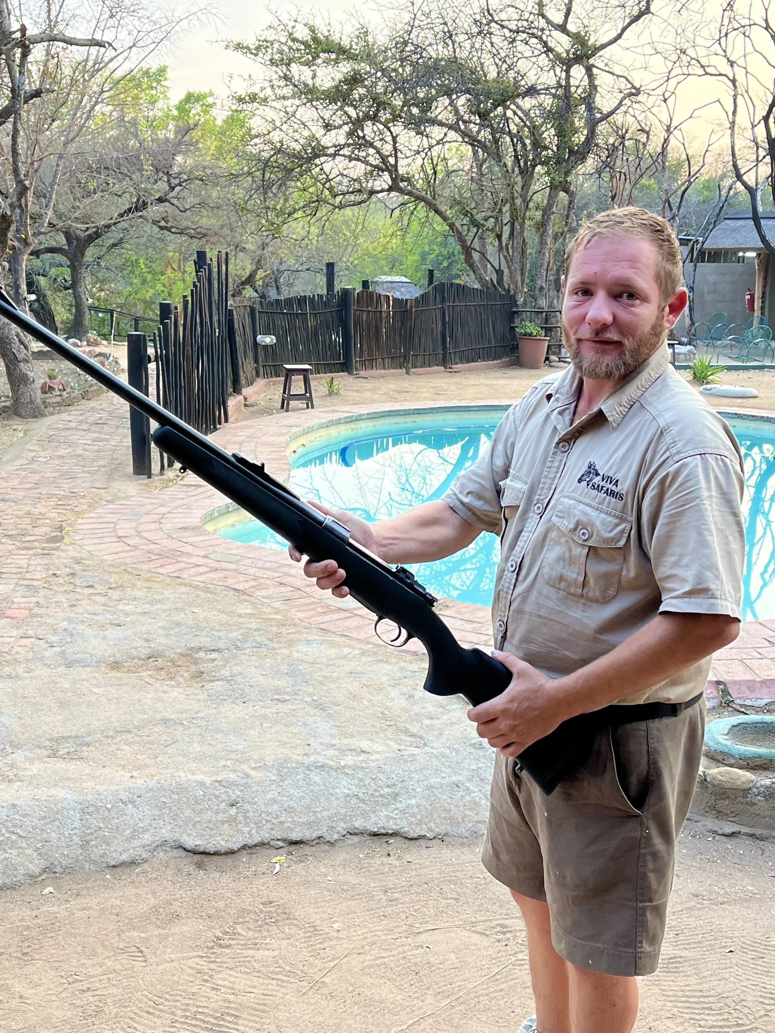 Man holding a rifle near a pool in a wooded outdoor area.
