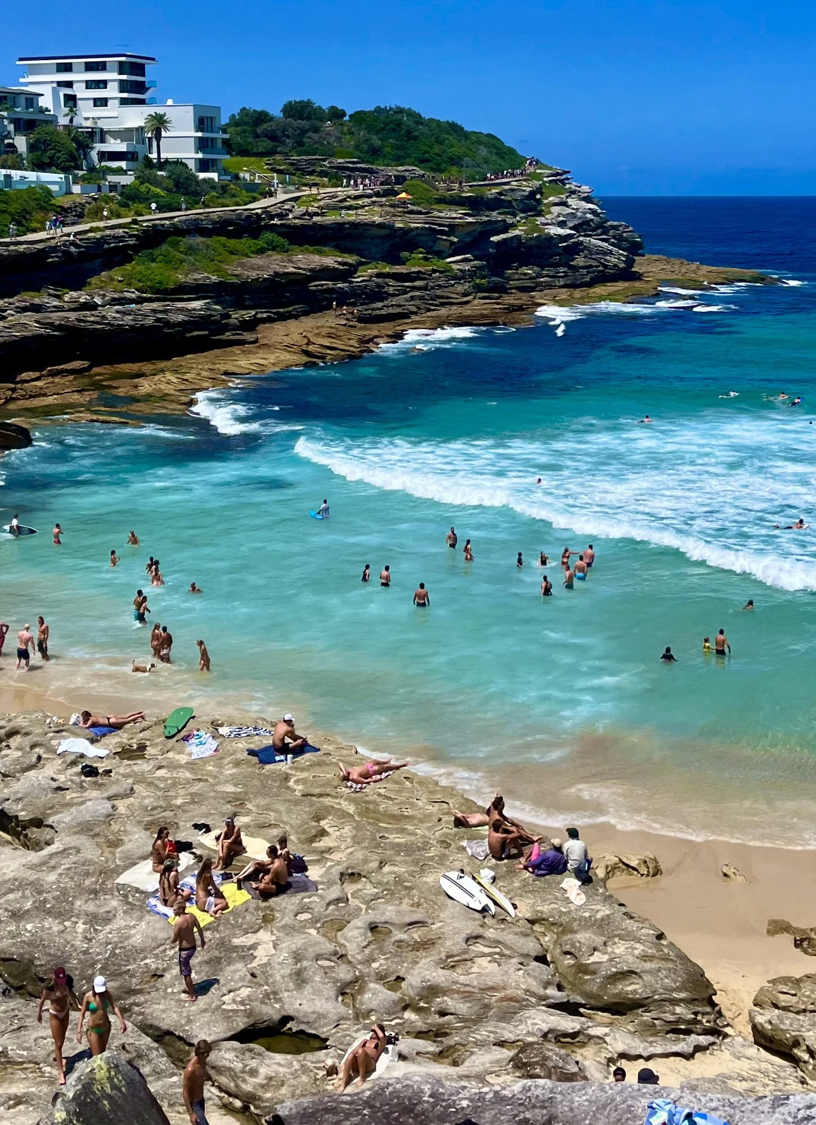 Beach scene with people swimming, sunbathing, and relaxing on rocks by the ocean.