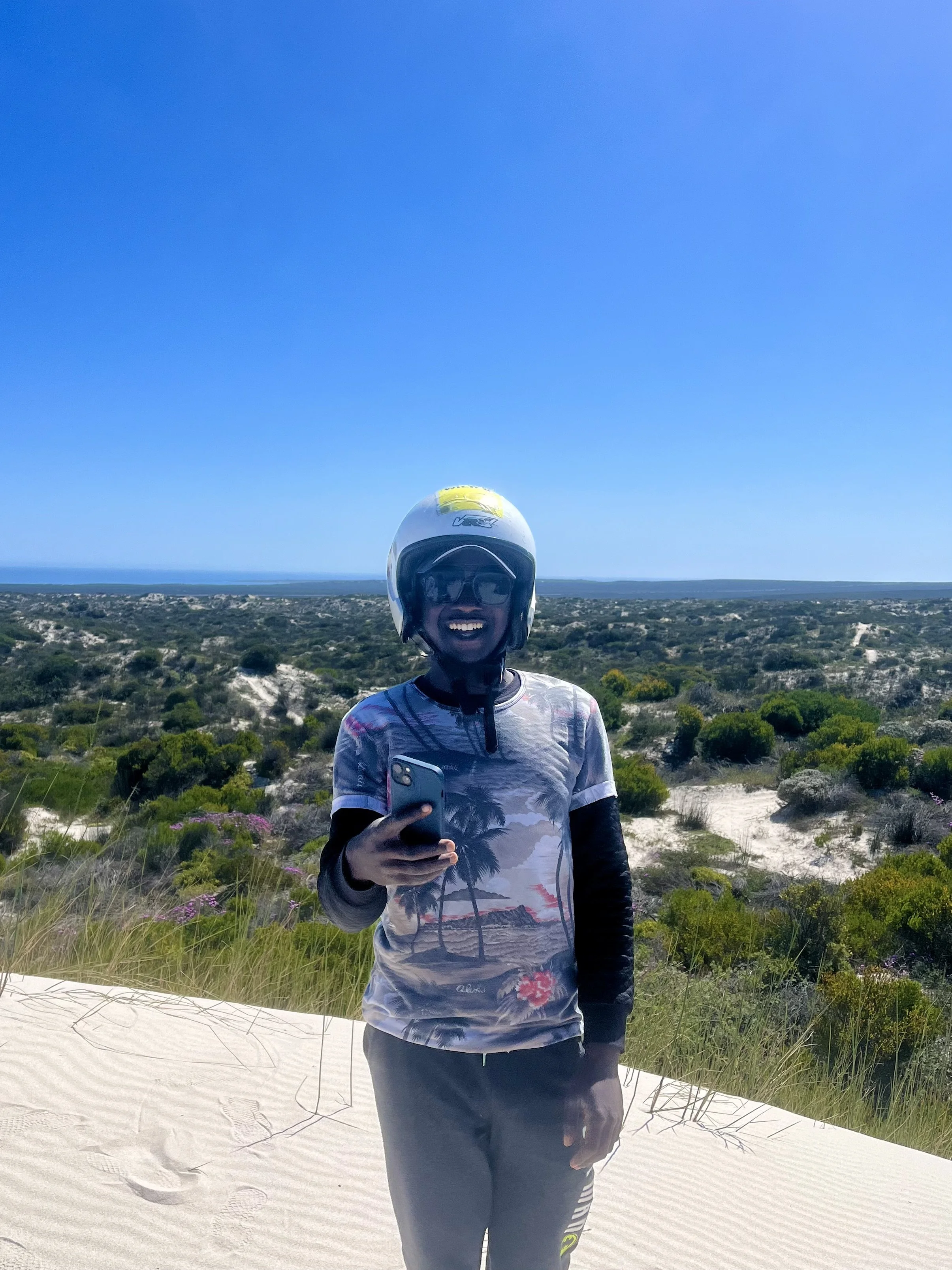 Person wearing a helmet and holding a smartphone, standing on a sandy area with vegetation and clear blue sky
