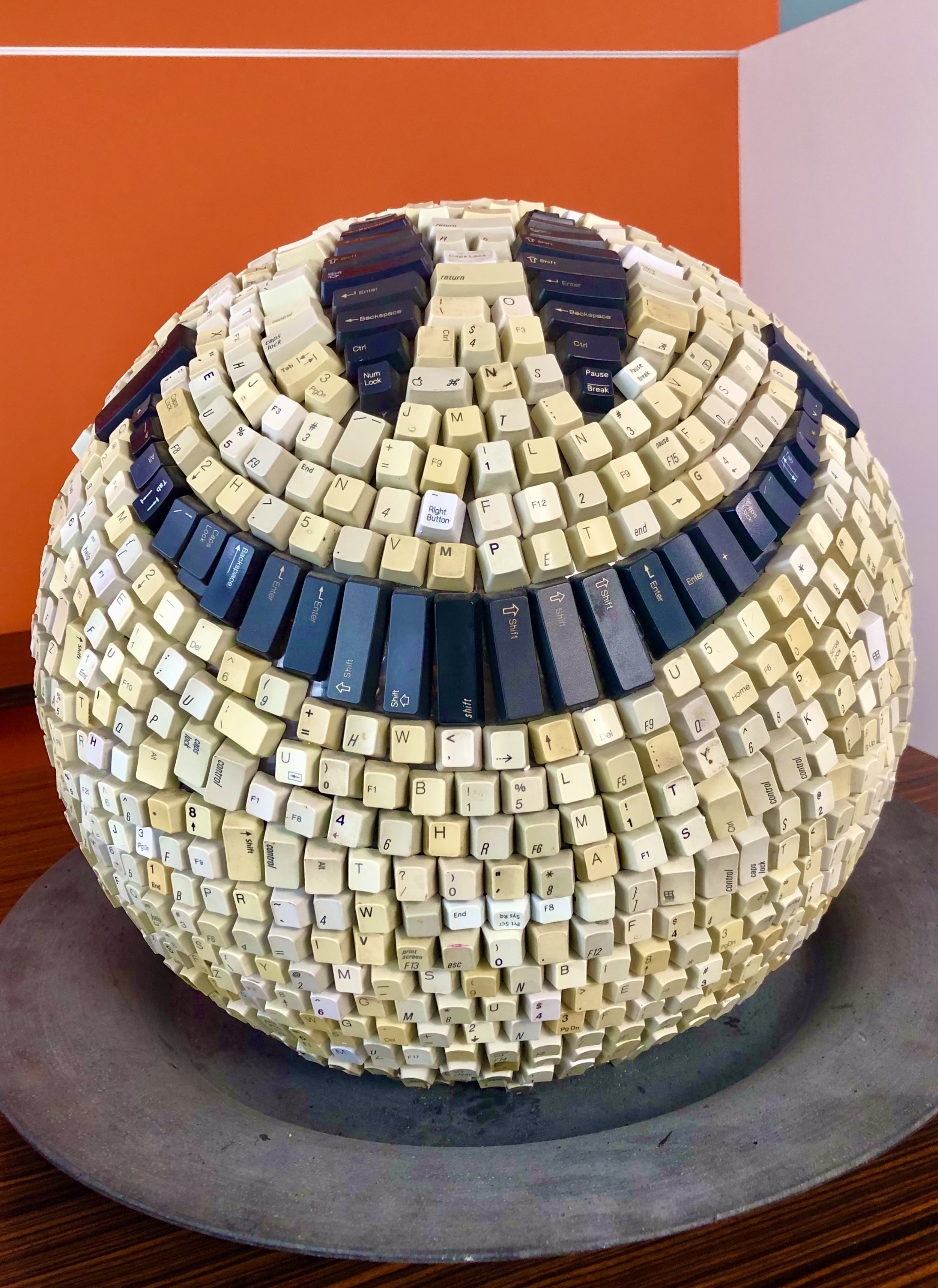 Sculpture of a sphere made from computer keyboard keys creating a face pattern on an orange and white background.