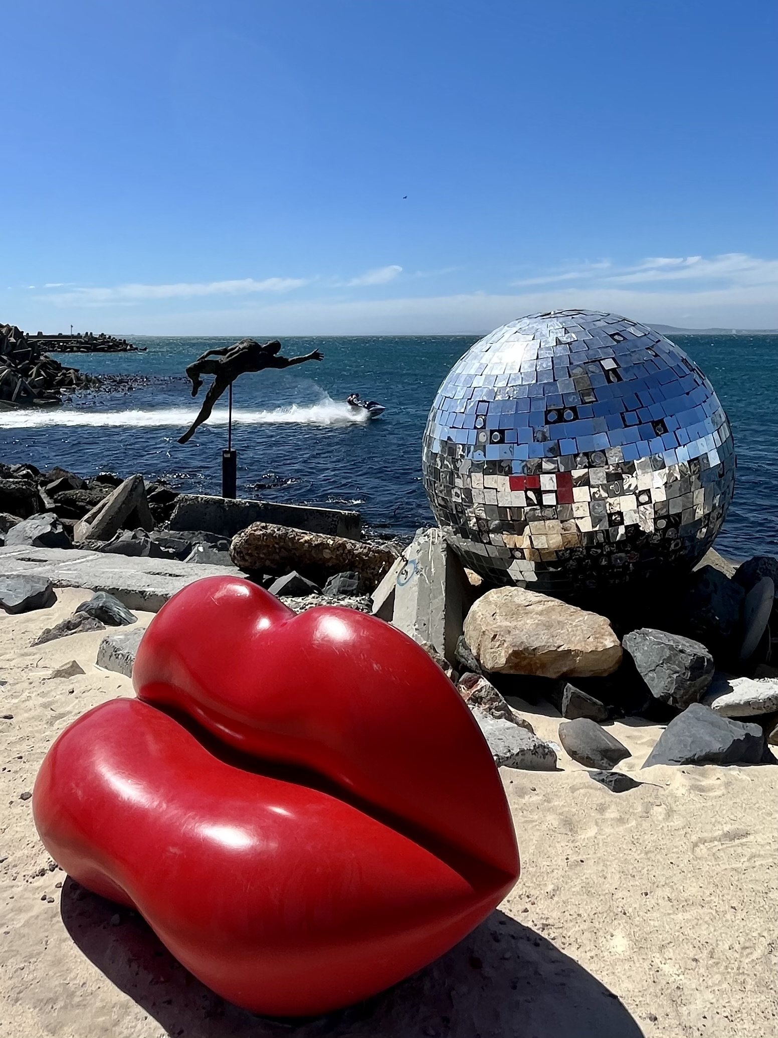 Large red lips sculpture on sandy beach, a disco ball on rocks, and a diving statue near water.